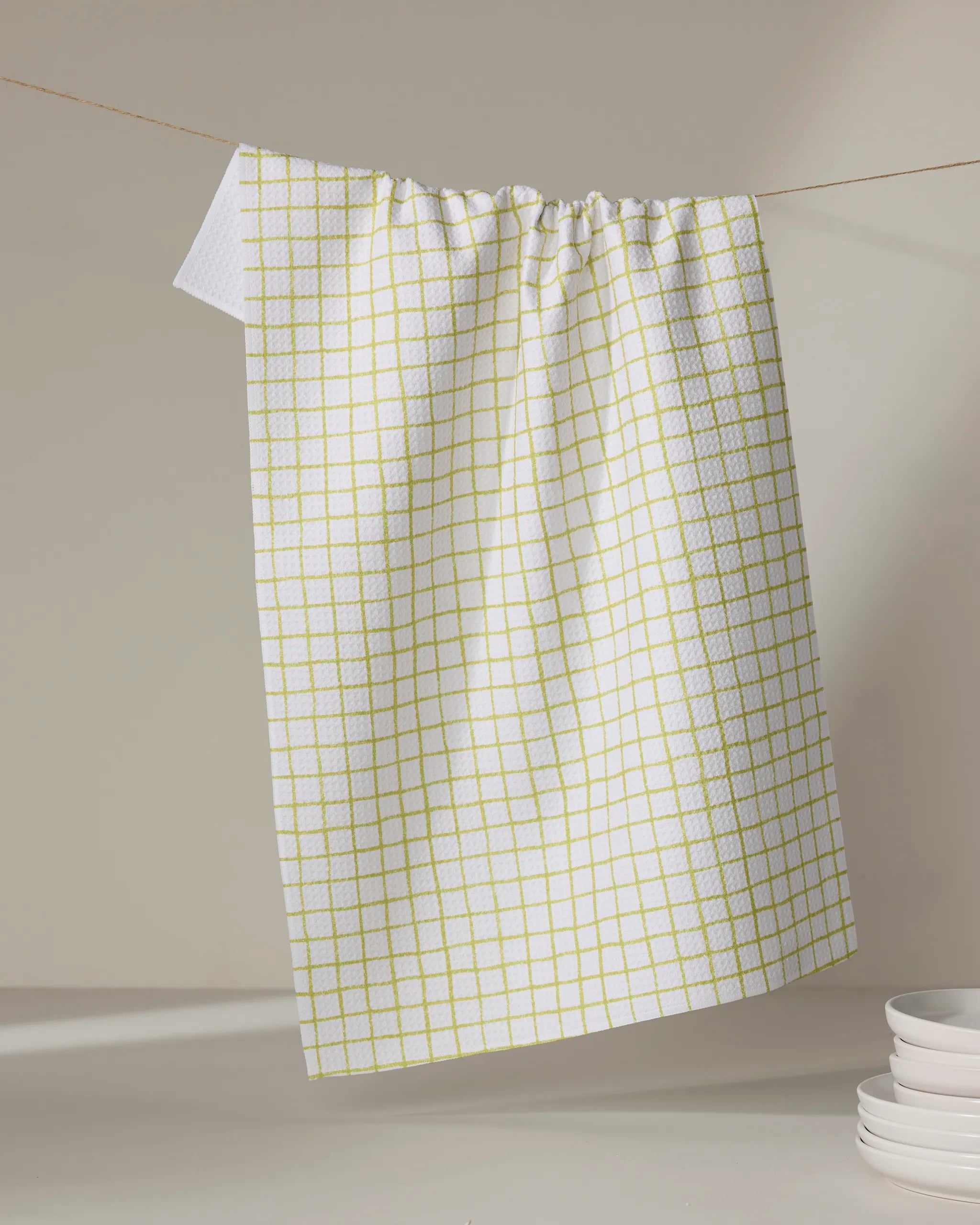 The On the Lawn kitchen towel by Kayla Ann, featuring a yellow grid pattern on white recycled fabric, hangs from a clothesline, casting a shadow on the background with a stack of white plates nearby.