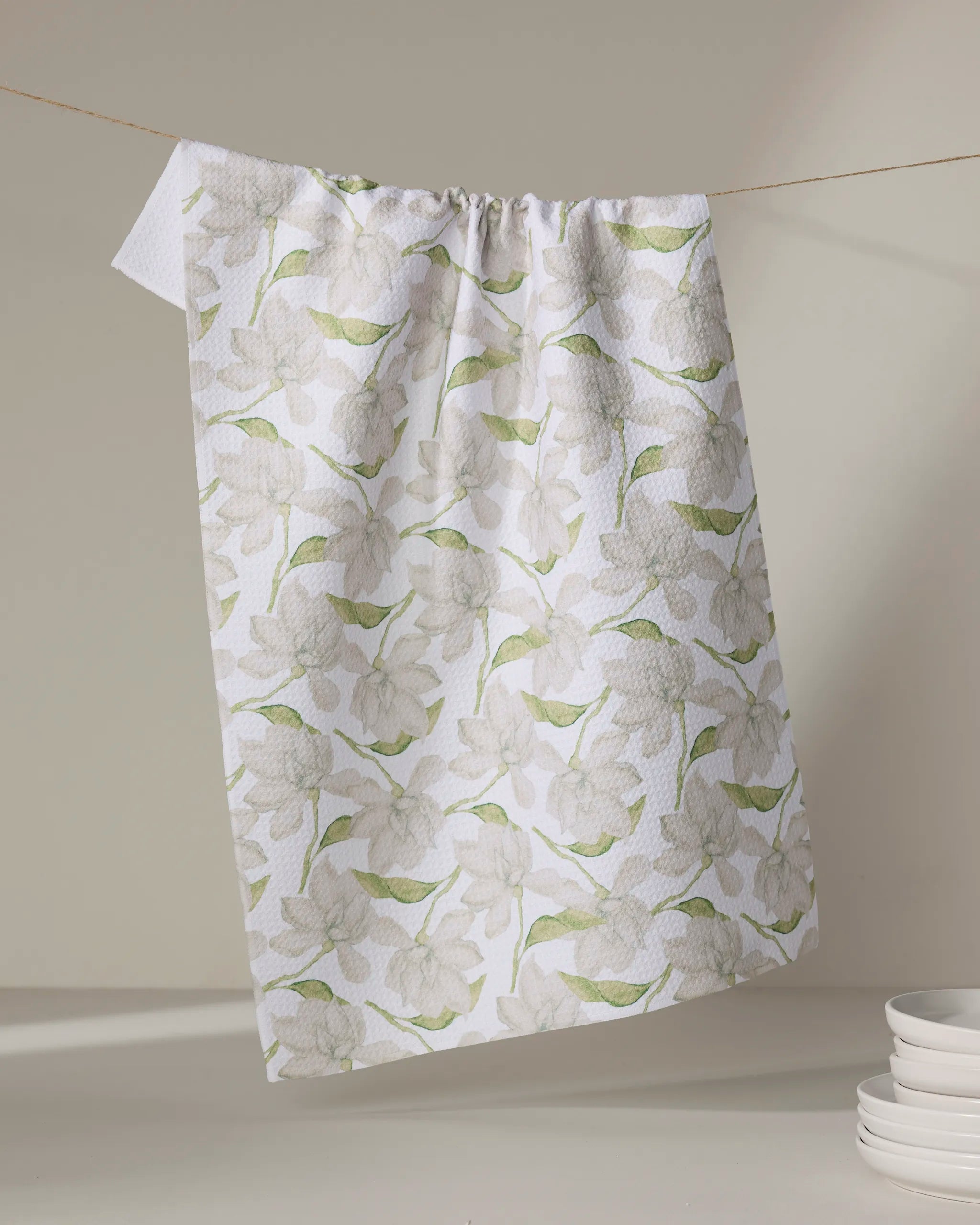 The "Once and a Floral" tea towel by Aubrey Fairchild, featuring a quick-drying white fabric with light green and beige floral patterns, hangs on a string above a stack of white plates against a softly lit neutral background.