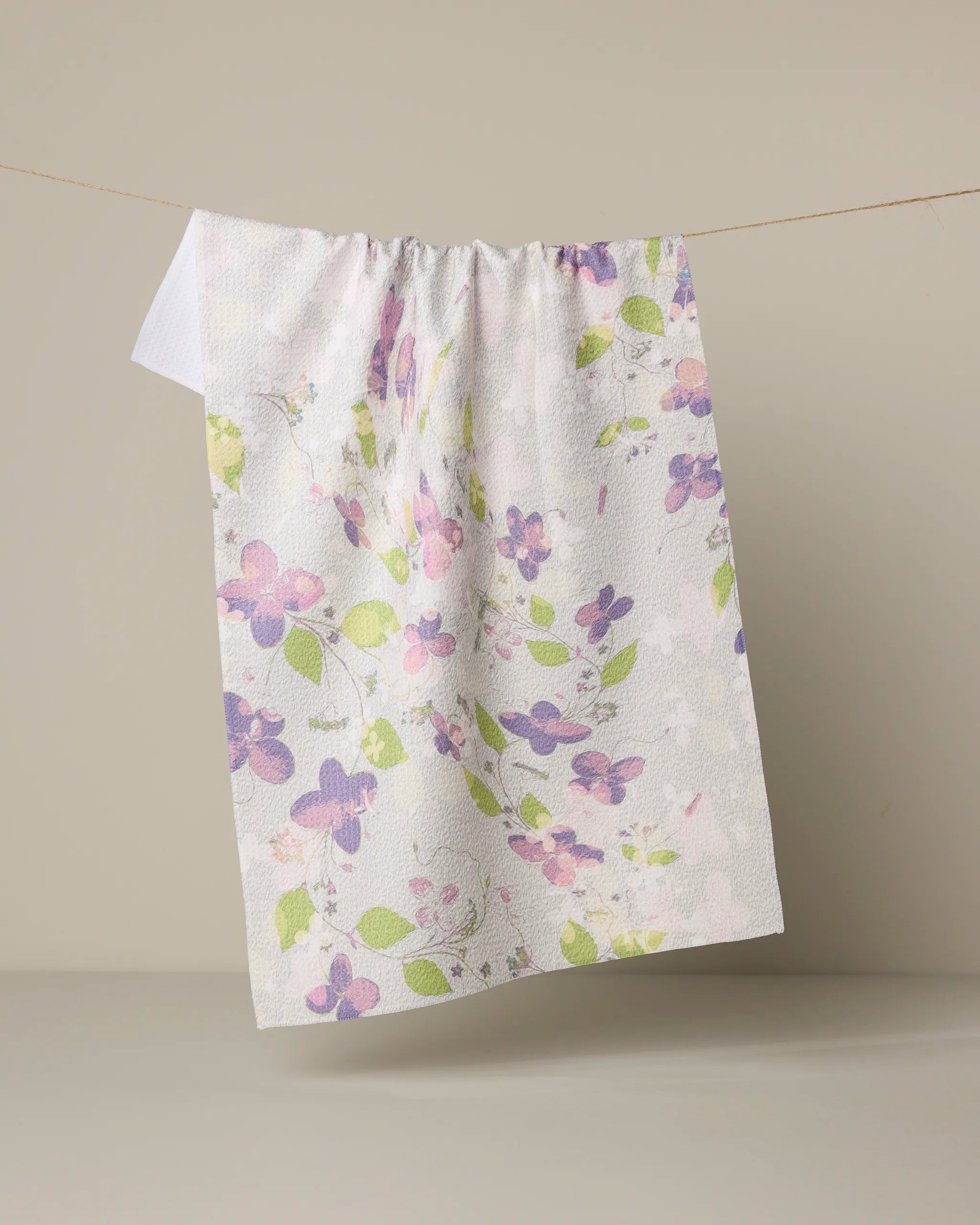 The Geometry Ophelia Garden tea towel, made from recycled materials, features a pastel floral pattern in purple, pink, and green on white and hangs against a neutral beige background.