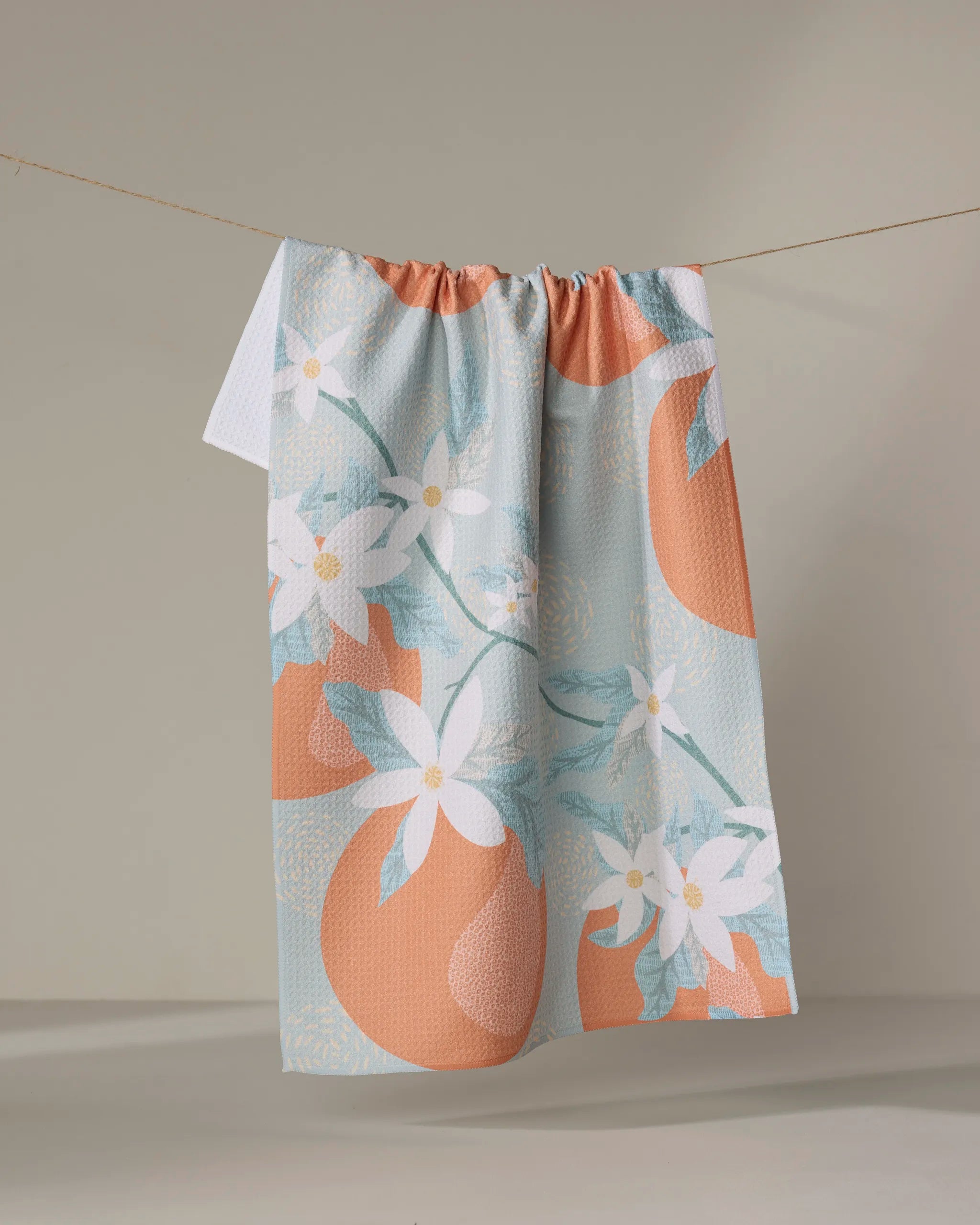 The Julianne Haness Orange Blossom tea towel, featuring large oranges, white blossoms, and green leaves on a pastel orange and white floral pattern, hangs on a thin string against a plain beige background.