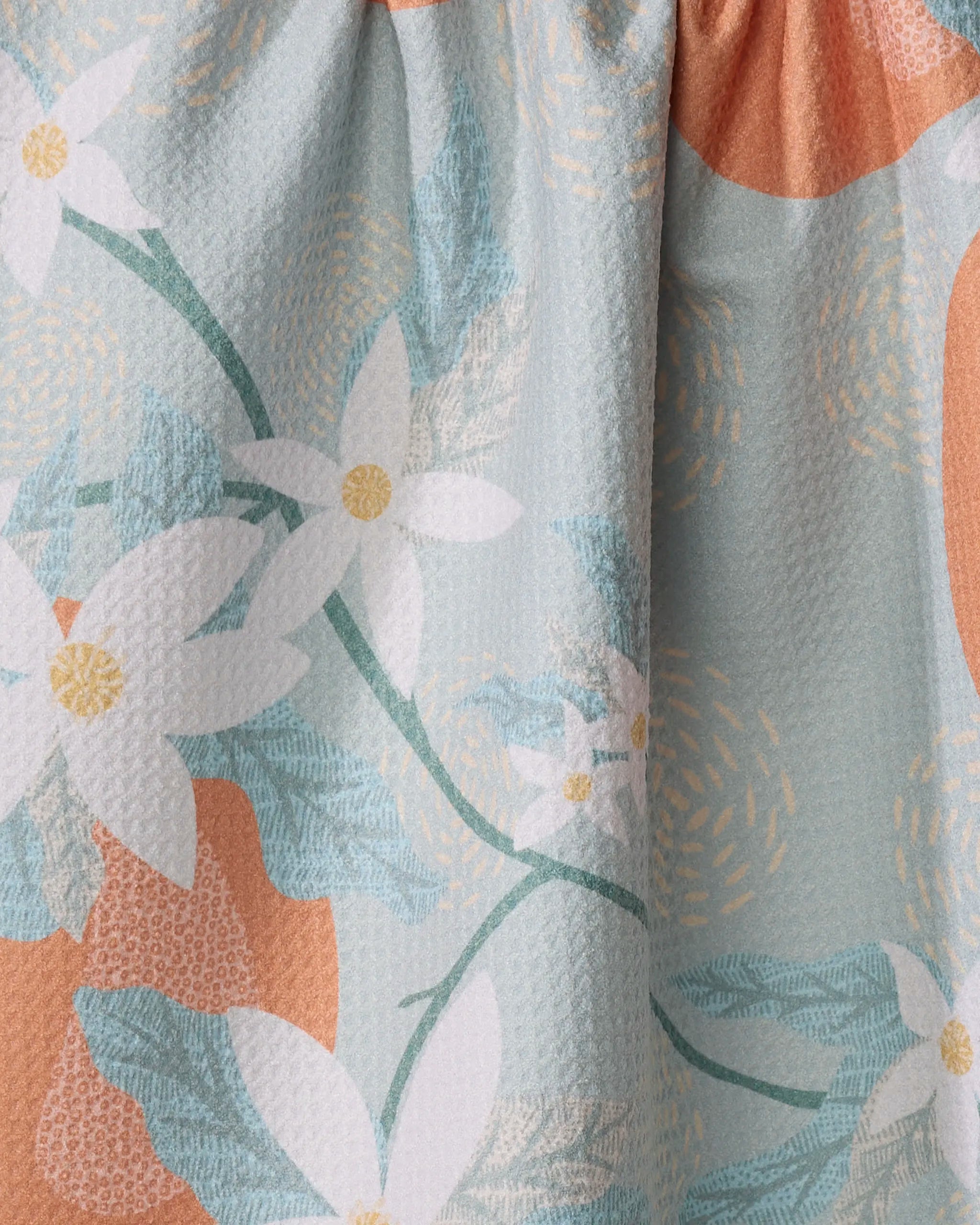 The Orange Blossom tea towel by Julianne Haness is light blue and peach with a textured, absorbent surface and gathered top. It features a floral pattern of white flowers with yellow centers and green leaves. Made from recycled materials.