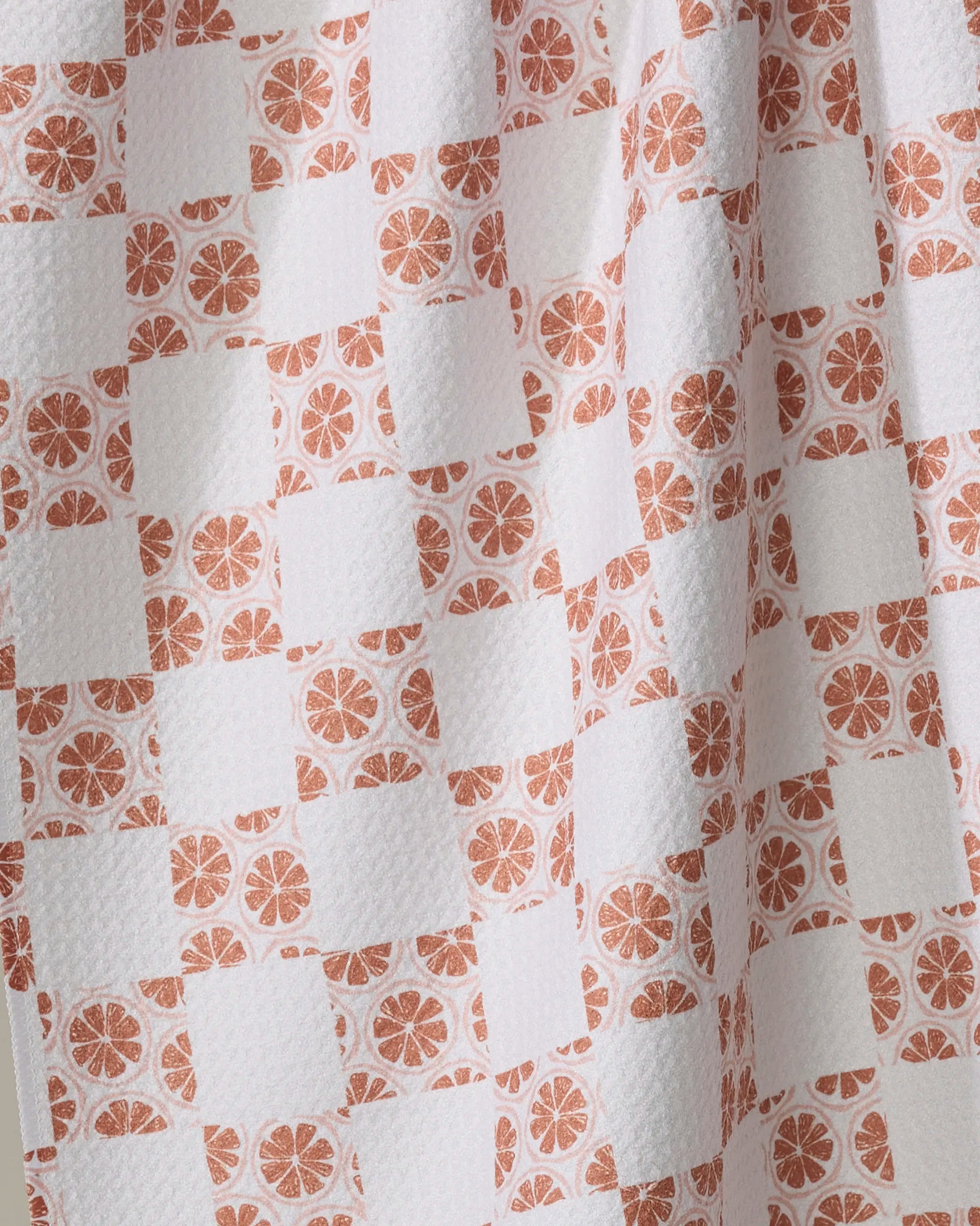 A close-up of Orange Checker by Teri the Butter shows textured white fabric made from recycled materials, with a checkered pattern of plain white squares and orange citrus slices. Soft draping creates gentle folds and shadows—ideal for a tea towel.