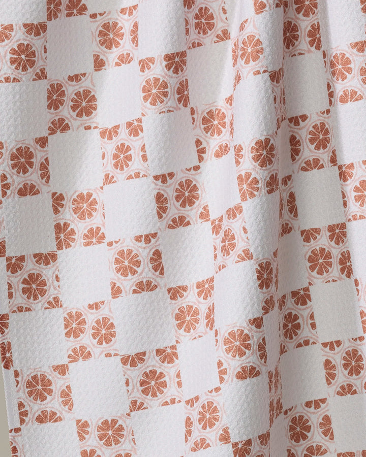 A close-up of Orange Checker by Teri the Butter shows textured white fabric made from recycled materials, with a checkered pattern of plain white squares and orange citrus slices. Soft draping creates gentle folds and shadows—ideal for a tea towel.
