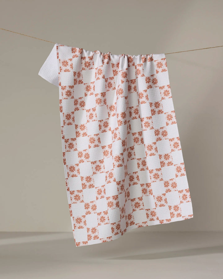 The Orange Checker tea towel by Teri the Butter, featuring a quick-drying checkered pattern with orange floral designs, hangs on a clothesline against a simple background.