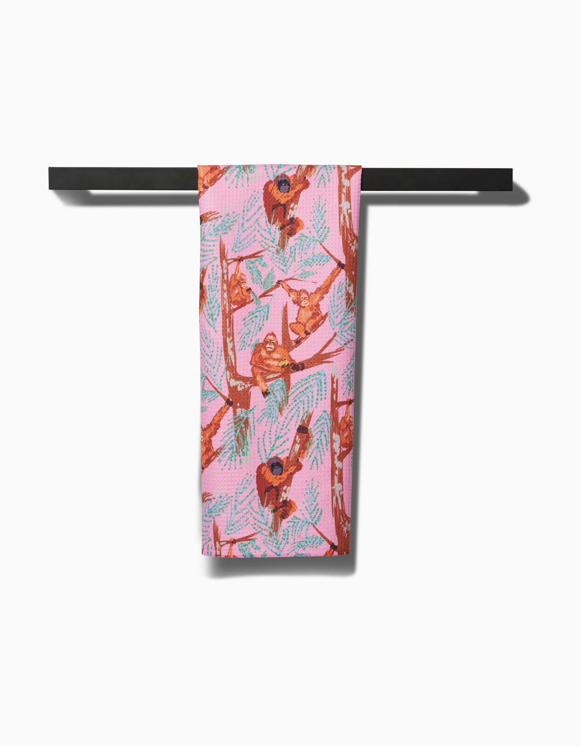 The Orangutan Clan tea towel by Elisabeth Wing, featuring colorful orangutans and green leaves on pink recycled fabric, hangs neatly over a black horizontal bar against a white background.