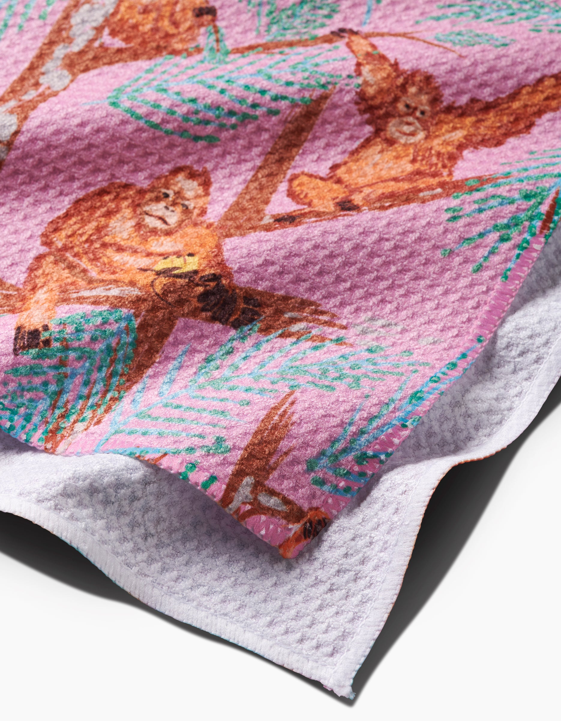 A close-up of the Orangutan Clan tea towel by Elisabeth Wing, showing orangutans in trees on a pink and green background. Made from recycled materials with a waffle weave and folded corner revealing the reverse side.