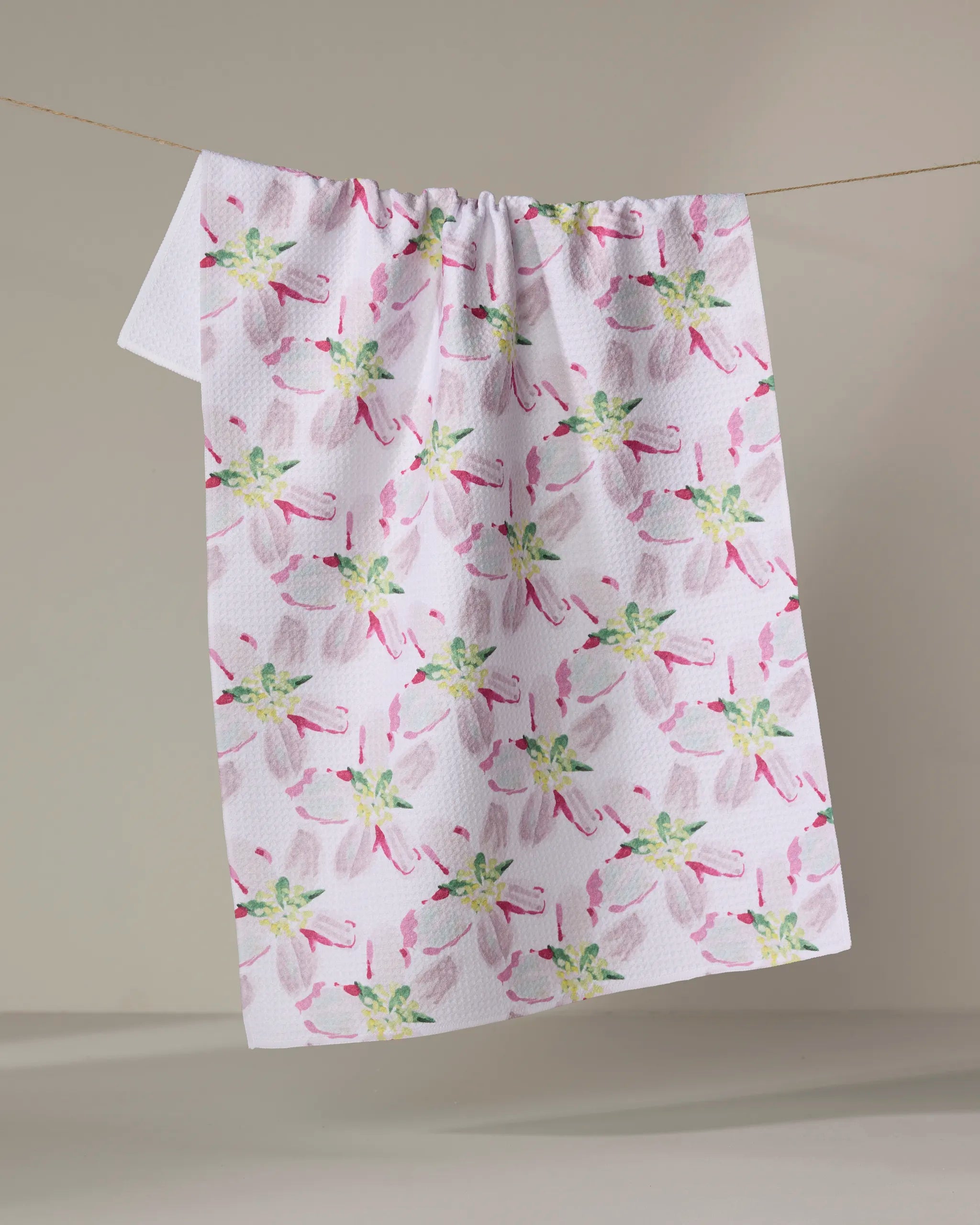 The Orchard in Bloom tea towel by Ania Zwara Art, ultra-absorbent and white with a pink, green, and pale purple floral pattern, hangs over a clothesline against a neutral background.