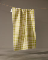 The Orchard Plaid Green tea towel by Geometry hangs vertically from a string against a gray background. Made from recycled materials, its lightweight fabric is gathered at the top for an eco-friendly, stylish touch.