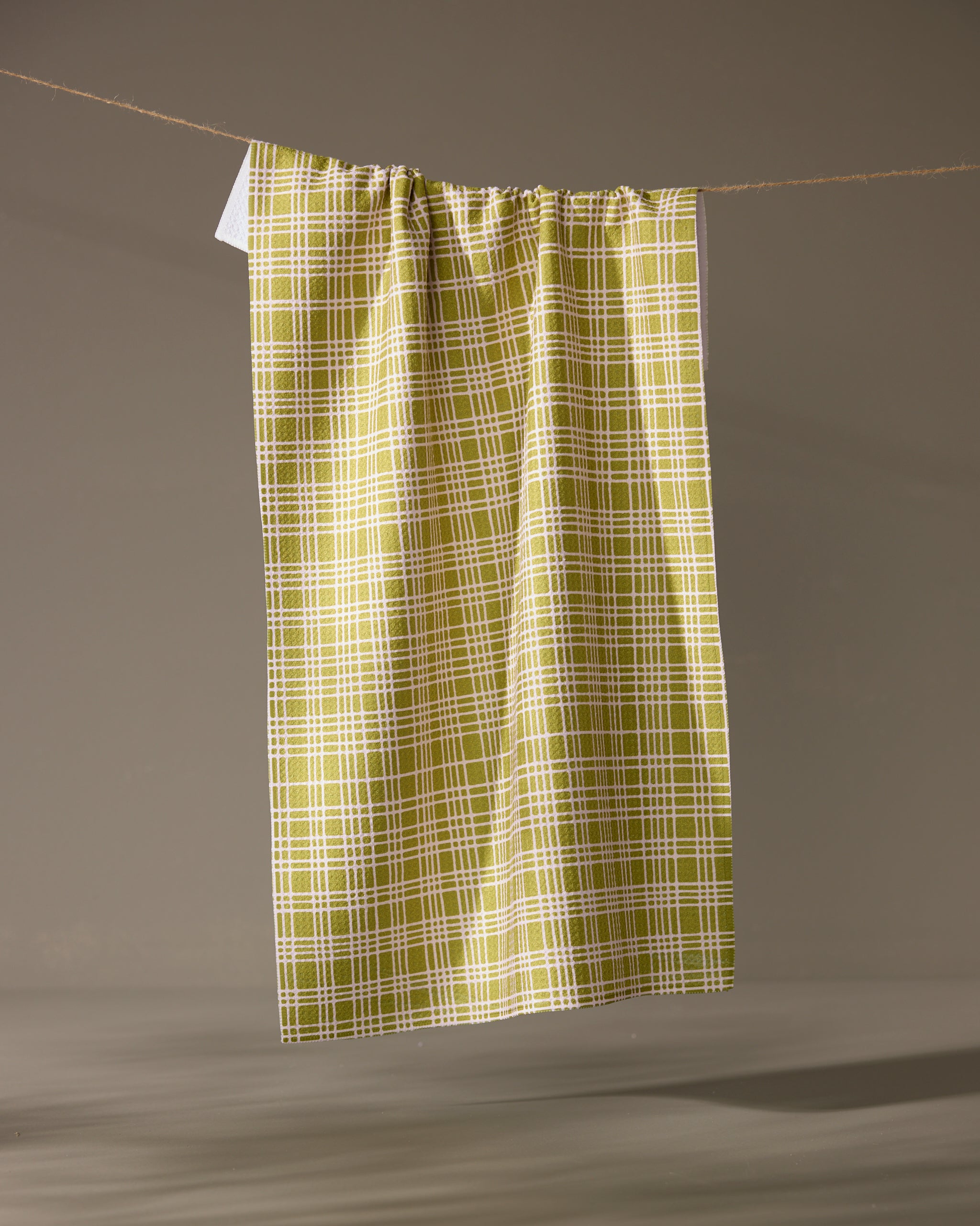 The Orchard Plaid Green tea towel by Geometry hangs vertically from a string against a gray background. Made from recycled materials, its lightweight fabric is gathered at the top for an eco-friendly, stylish touch.