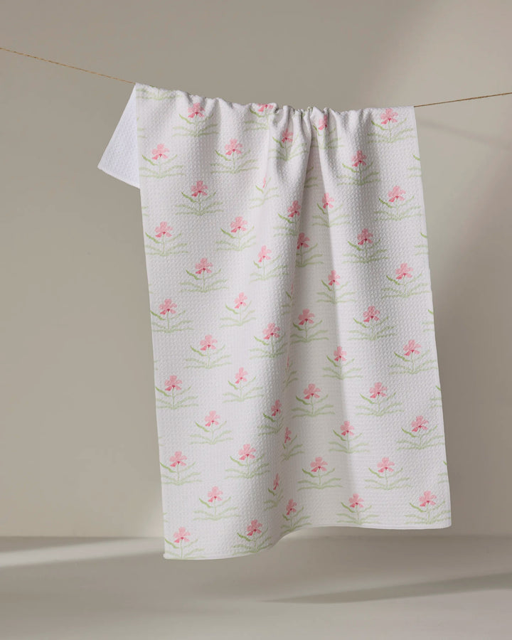 The Orchids In Bloom tea towel by Laura Prietto, crafted from recycled materials, displays pink flowers and green stems on white textured fabric as it hangs over a clothesline against a neutral background.
