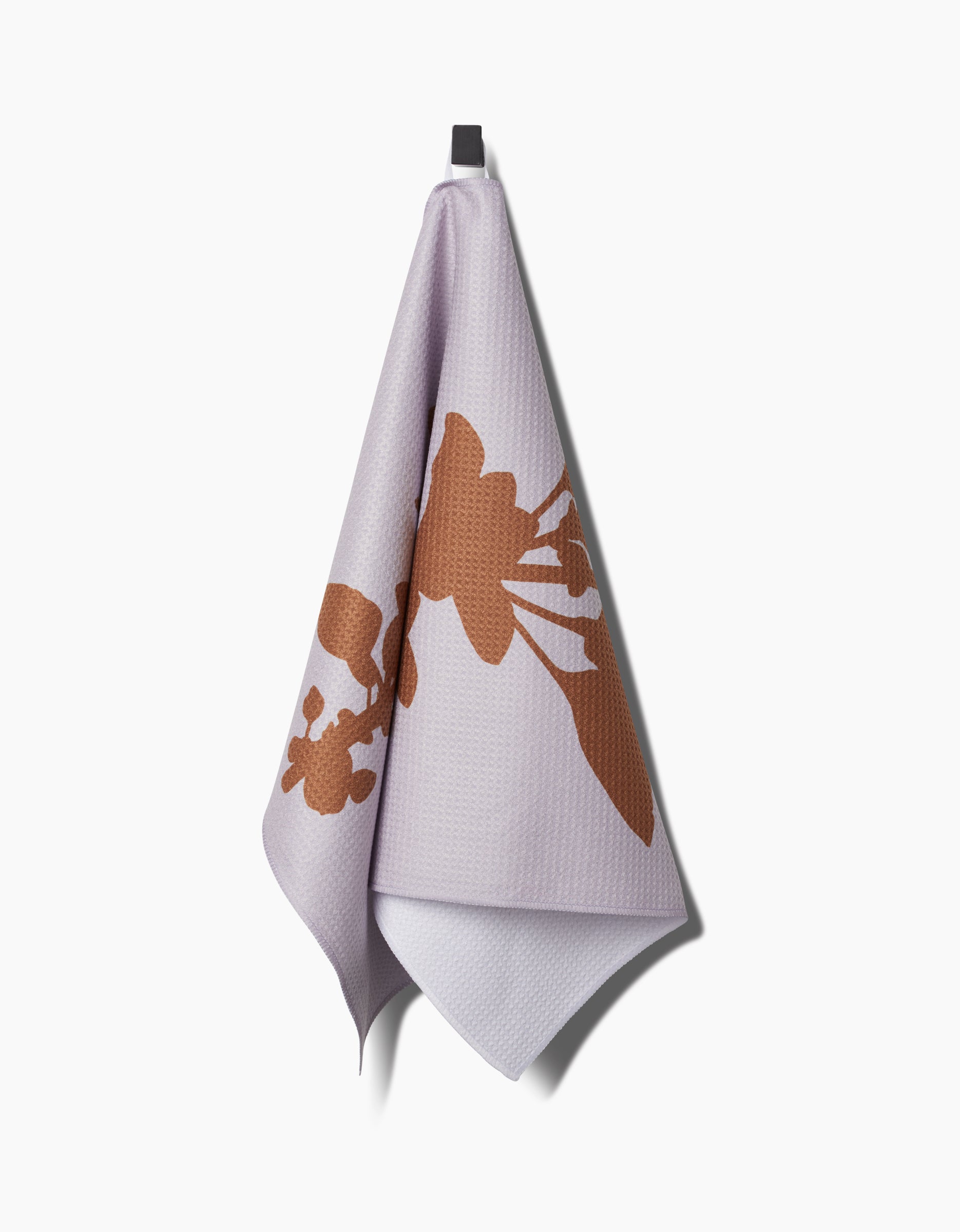 An Oregano Geoweave™ tea towel by PrintedPlant, featuring a large brown abstract floral pattern, hangs from a black hook against a white background.