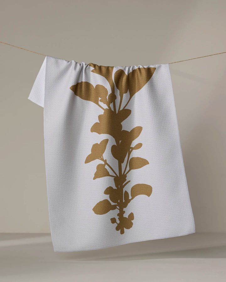 PrintedPlant’s “Oregano” is a white textured tea towel featuring a bold gold botanical silhouette, made from recycled materials and displayed hanging on a thin string against a neutral background.