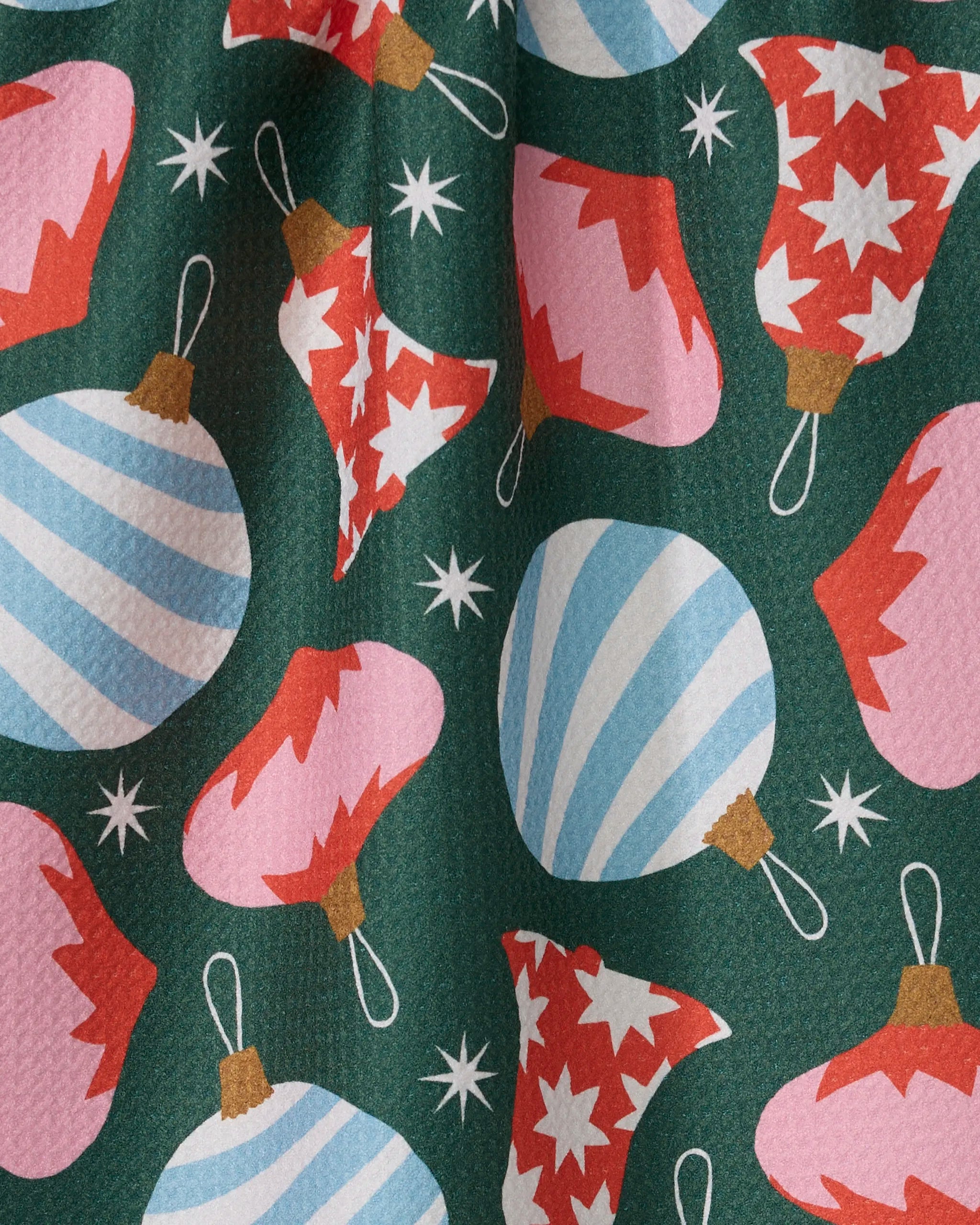The Ornamenta kitchen towel by Geometry features a festive recycled design with blue stripes, pink Christmas ornaments, red and white starbursts, and scattered white stars on a dark green background.