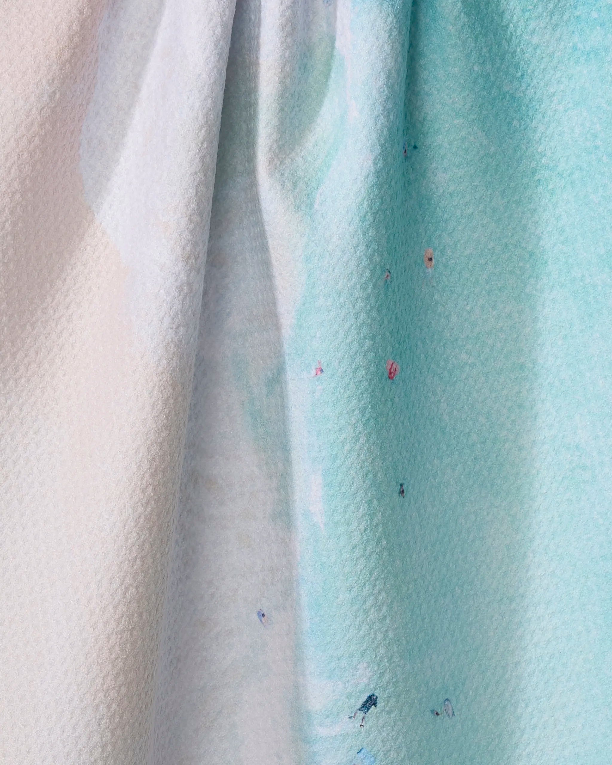 A close-up of Kait Hanson's "Out to Sea" fabric showcases soft pastel blue and white hues from recycled materials, accented with small colorful paint splatters and gentle folds.