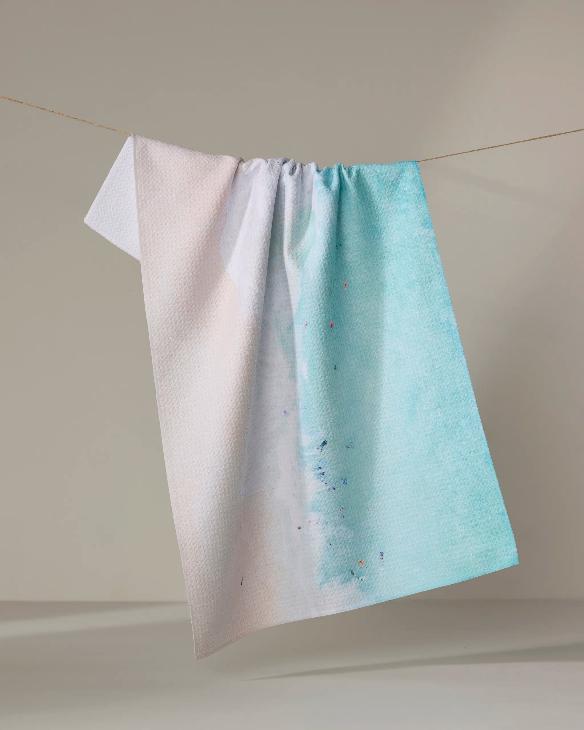 The "Out to Sea" tea towel by Kait Hanson, crafted from recycled materials, features a textured design with a white-to-blue gradient and hangs over a clothesline against a neutral background.