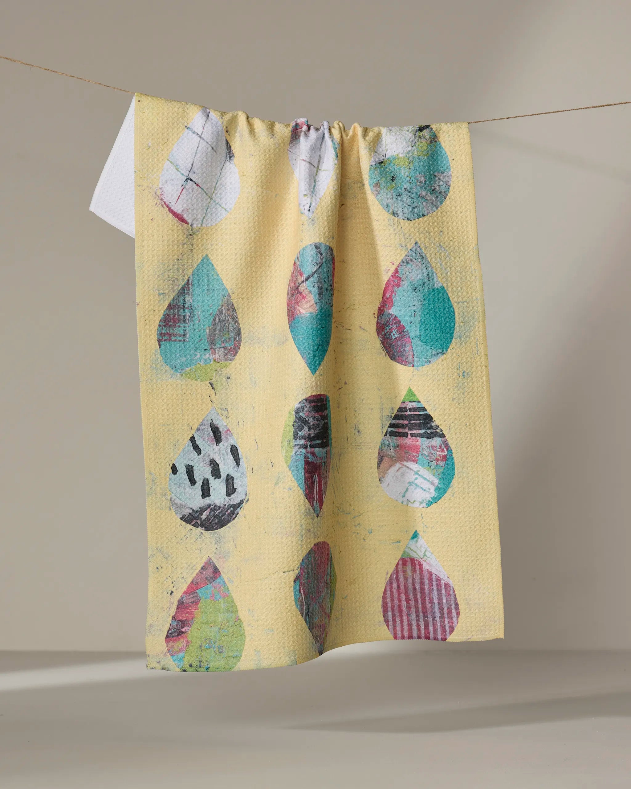The Outer Limits recycled kitchen towel by Katherine Berkeley features colorful abstract raindrop shapes in blue, teal, pink, and white on yellow, hanging against a neutral background.