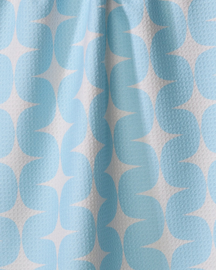 A close-up of Mindfulnice's "Over the River" tea towel, made from recycled materials, shows its textured fabric and a repeating pattern of light blue S-shaped curves on a white background, slightly gathered at the center.