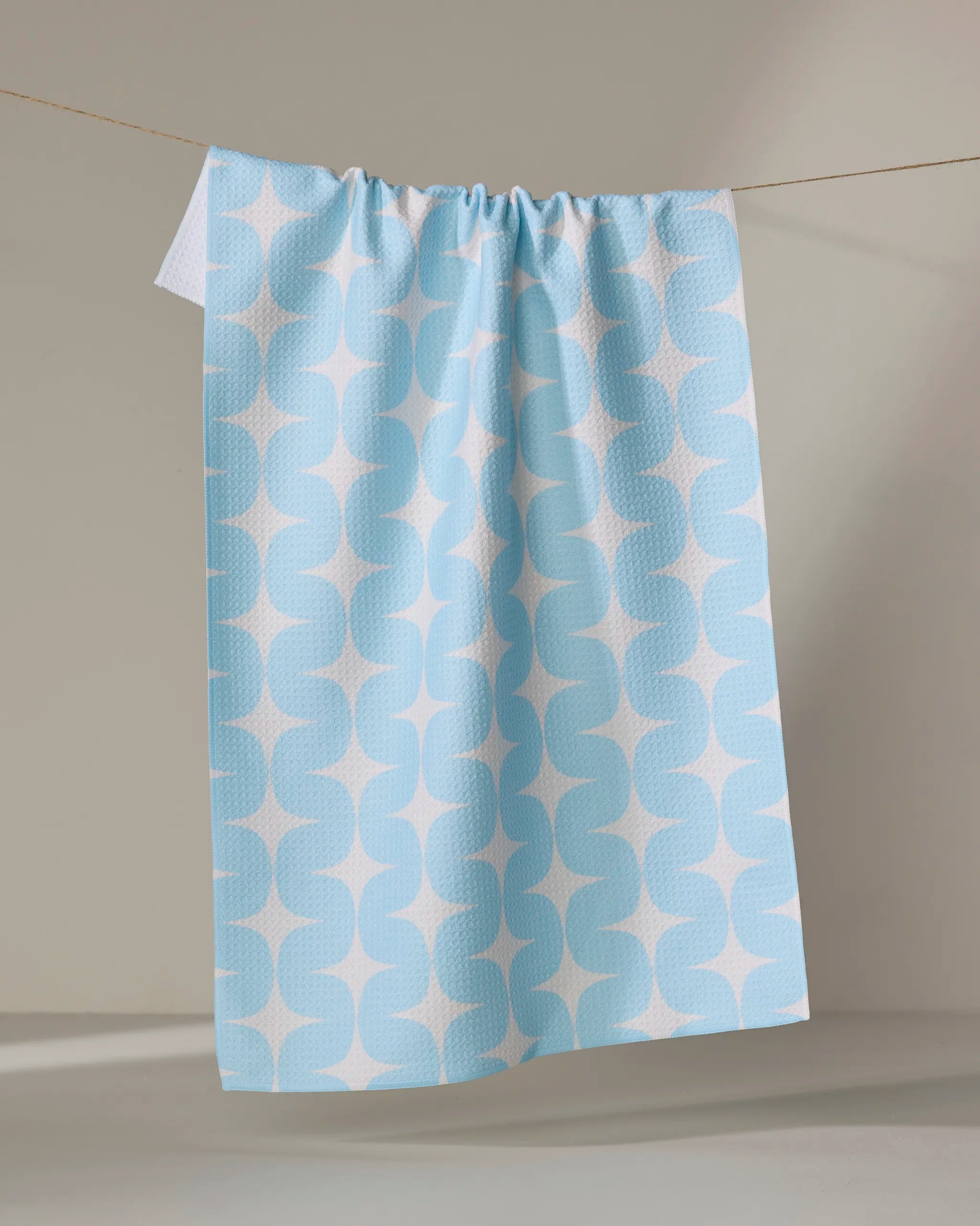 The Mindfulnice "Over the River" kitchen towel, made from recycled materials and featuring a light blue base with a white geometric pattern, hangs on a thin clothesline against a neutral background.