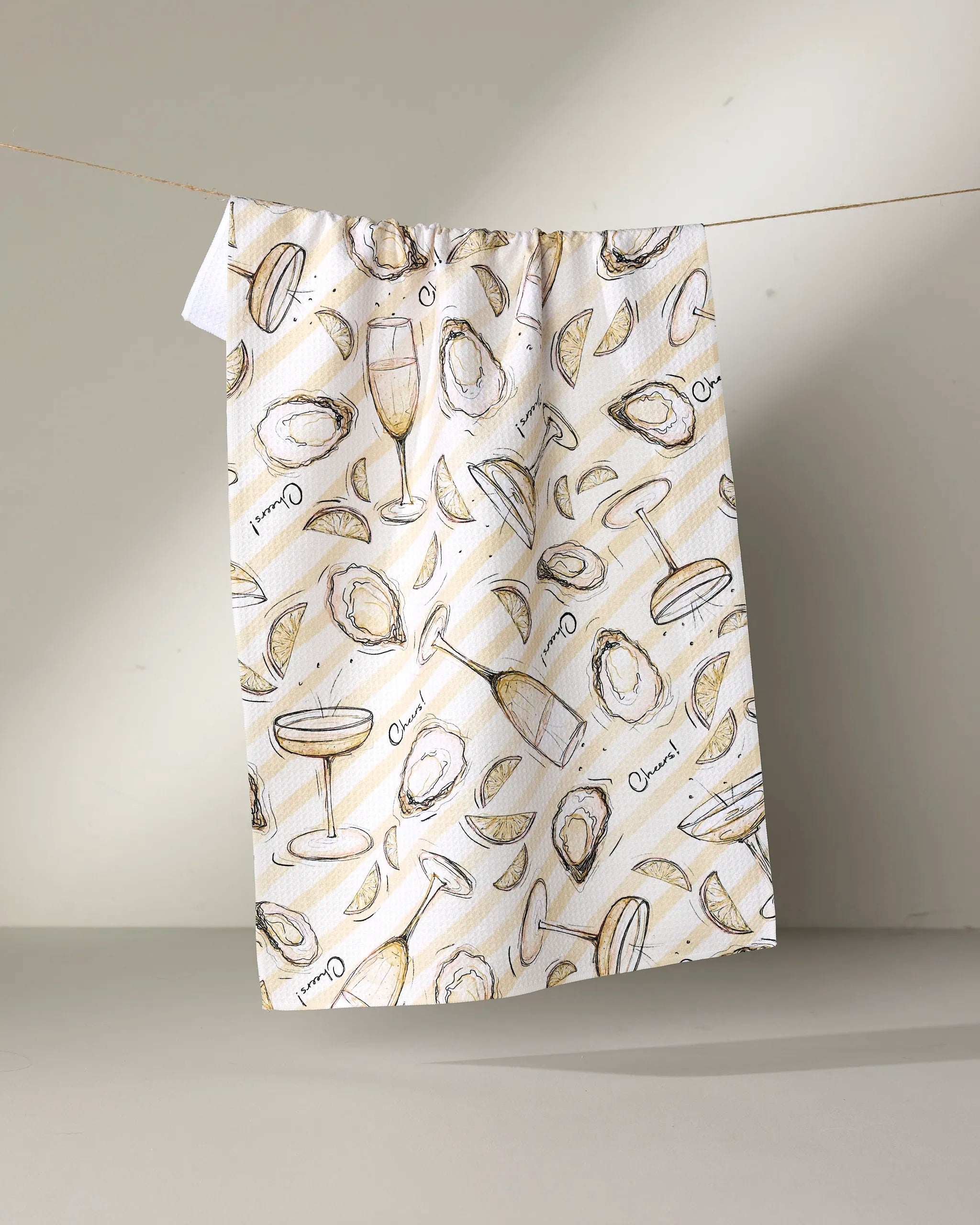 The Geometry "Oysters and Champagne" recycled kitchen towel hangs on a line, featuring oysters, lemon wedges, and champagne glasses in beige, gold, and black on a light background.