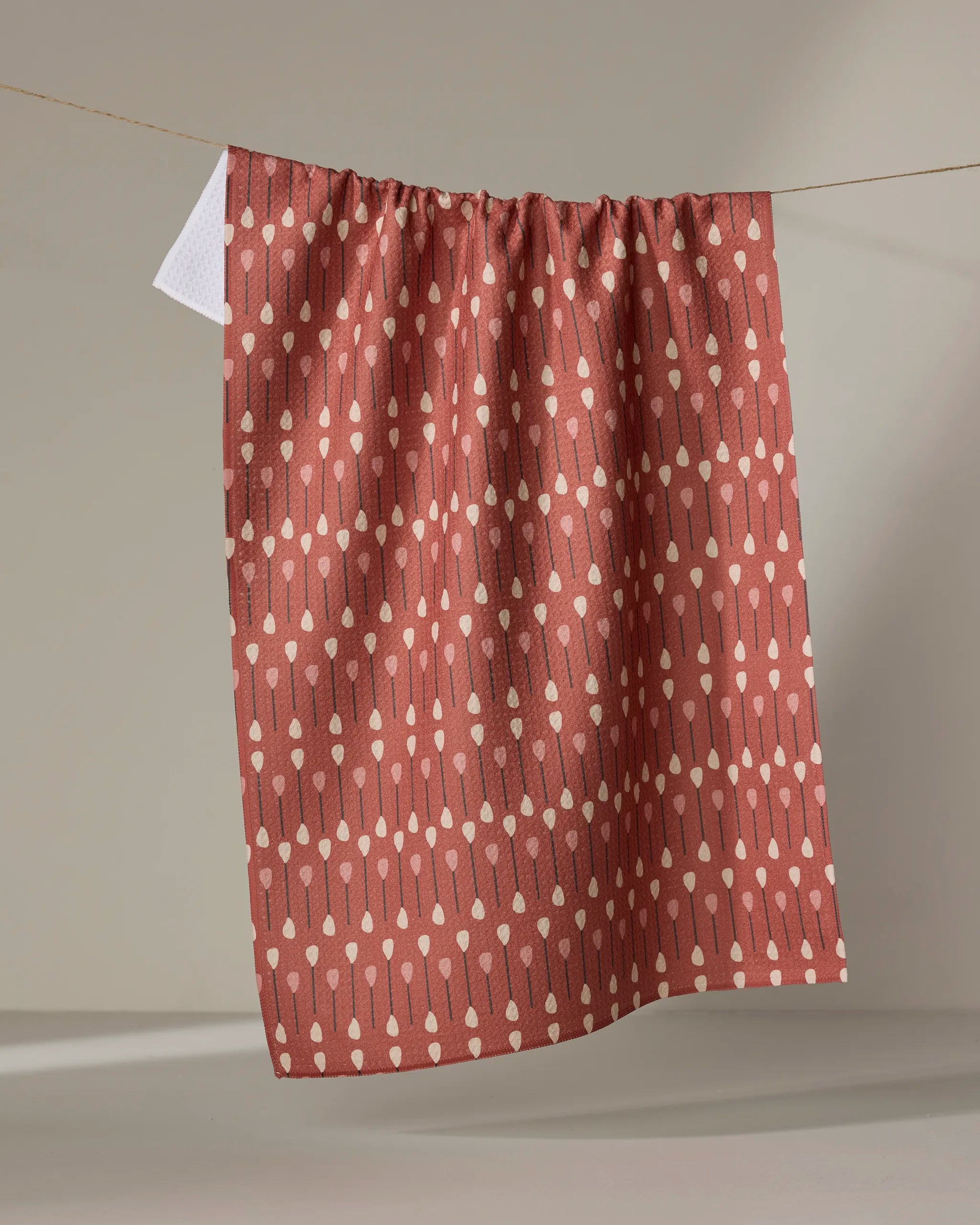 The "Paddles and Oars" tea towel by Coit Creative, made from recycled materials, features a reddish-brown hue with vertical cream dots and lines, and is shown hanging against a neutral gray background.