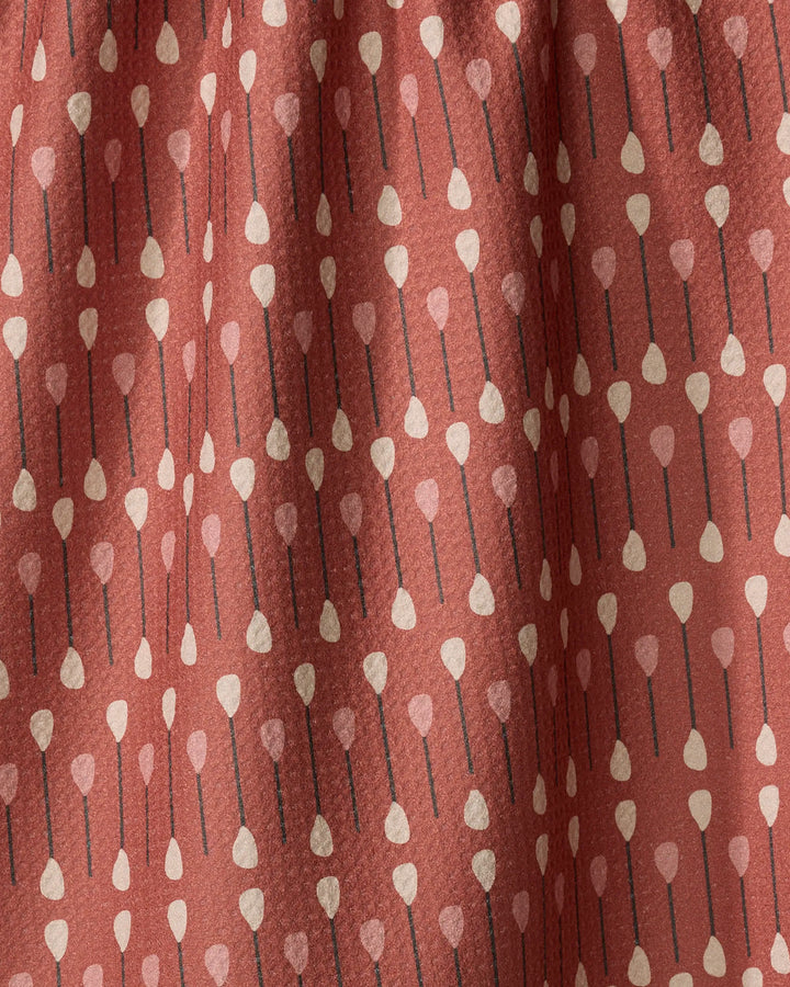 The Coit Creative "Paddles and Oars" tea towel features a rust color and vertical rows of stylized paddles with beige tips, black shafts, and light pink heart-shaped ends. Made from quick-drying recycled materials and shown with gentle folds.