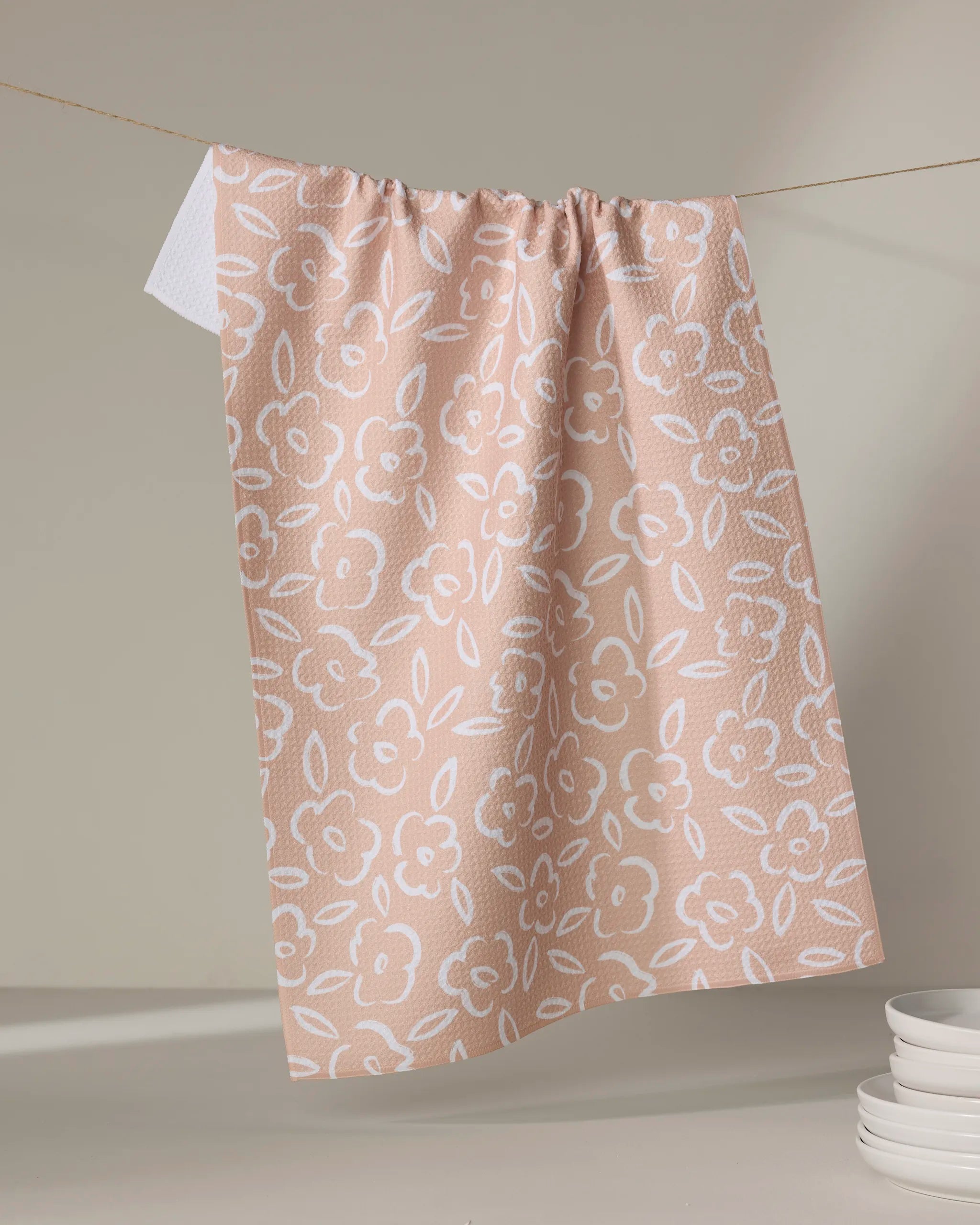 The Painted Bloom dish towel by Hufton Studio, featuring a pink and white floral design, hangs on a clothesline with a neutral backdrop and a stack of white plates in the lower right corner.