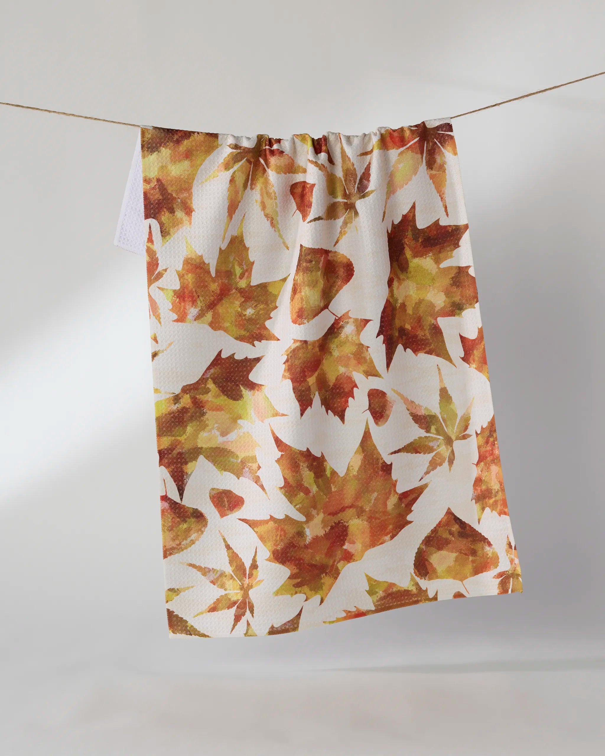 The Geometry Painted Fall dish towel, white and absorbent with orange, yellow, and brown autumn leaves, hangs on a string against a plain light background.