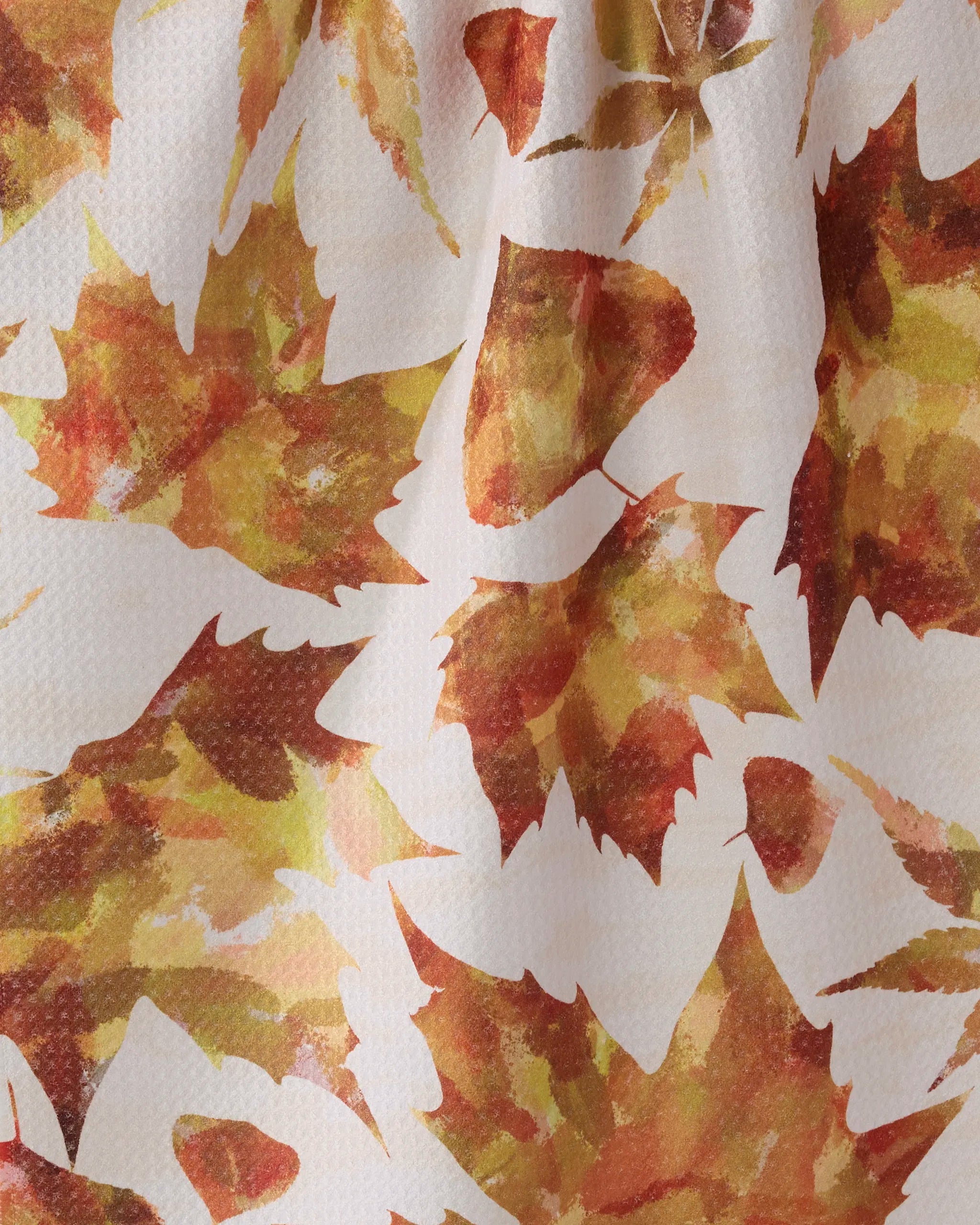 A close-up of Geometry’s Painted Fall tea towel, featuring an absorbent, slightly wrinkled fabric with autumn leaves in brown, yellow, and orange on a white background.