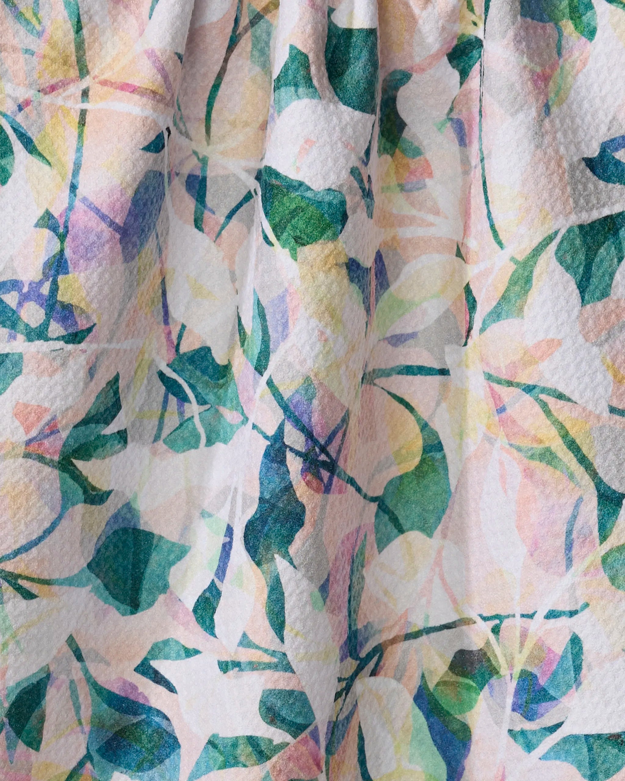 A close-up of the Painted Foliage kitchen towel by Rachel Parker, featuring a colorful abstract leaf and branch pattern in green, pink, yellow, and blue on textured fabric made from recycled materials. The fabric is softly gathered.