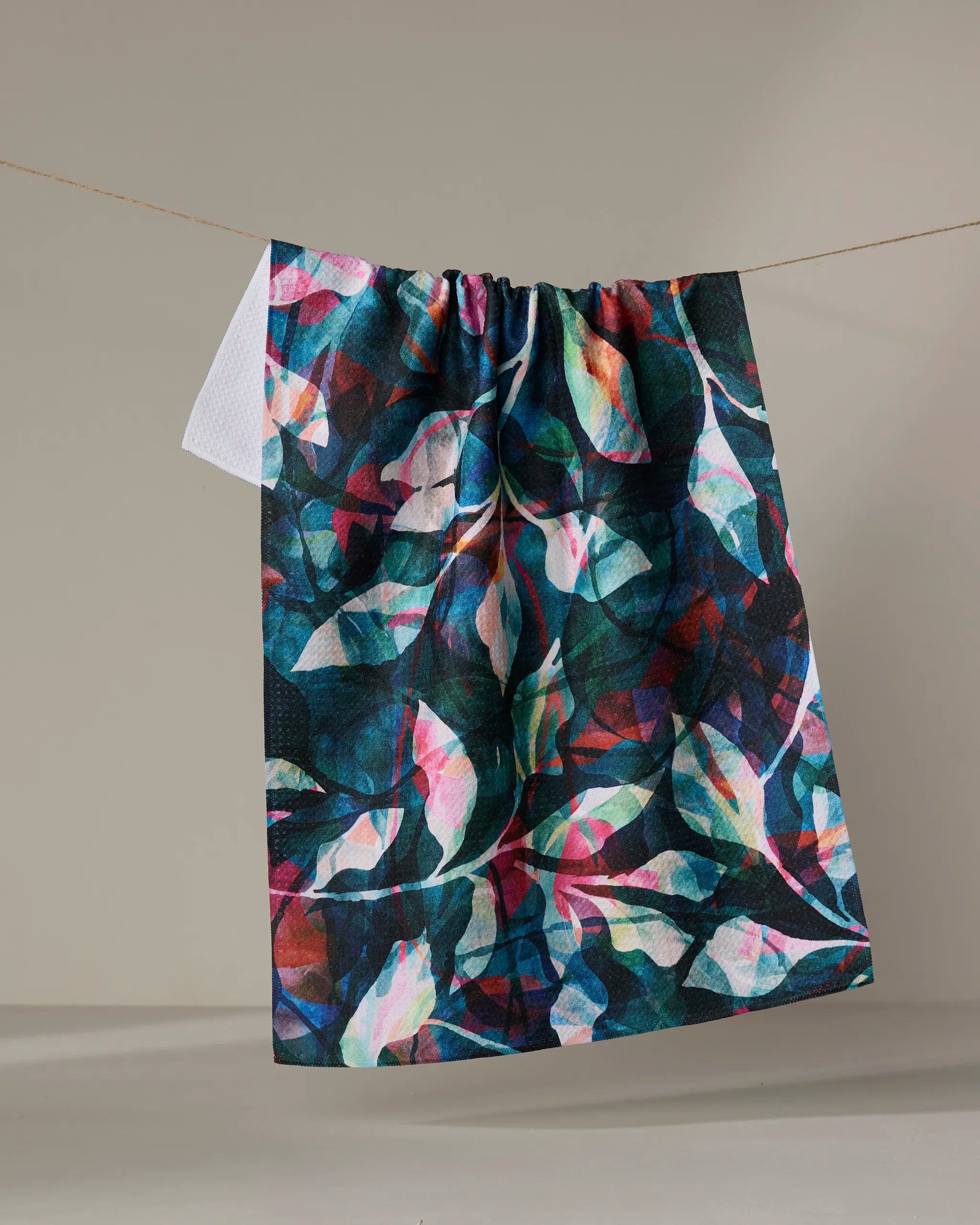 The Rachel Parker Painted Foliage Teal tea towel, featuring an abstract leaf design in blue, green, pink, and white, is displayed hanging on a clothesline against a simple background.