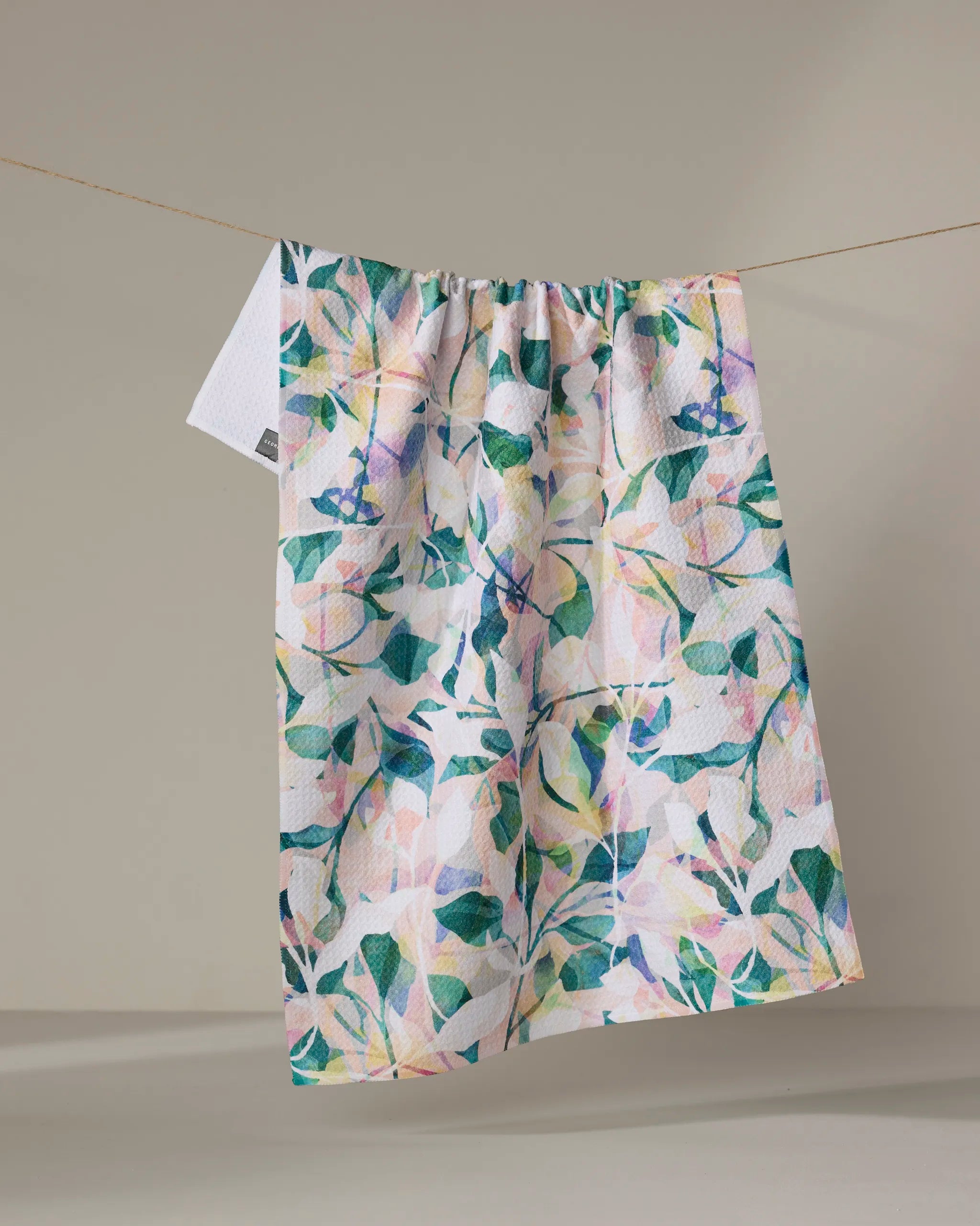 The "Painted Foliage" tea towel by Rachel Parker, crafted from recycled materials and featuring a leafy floral design, hangs on a clothesline against a neutral background.