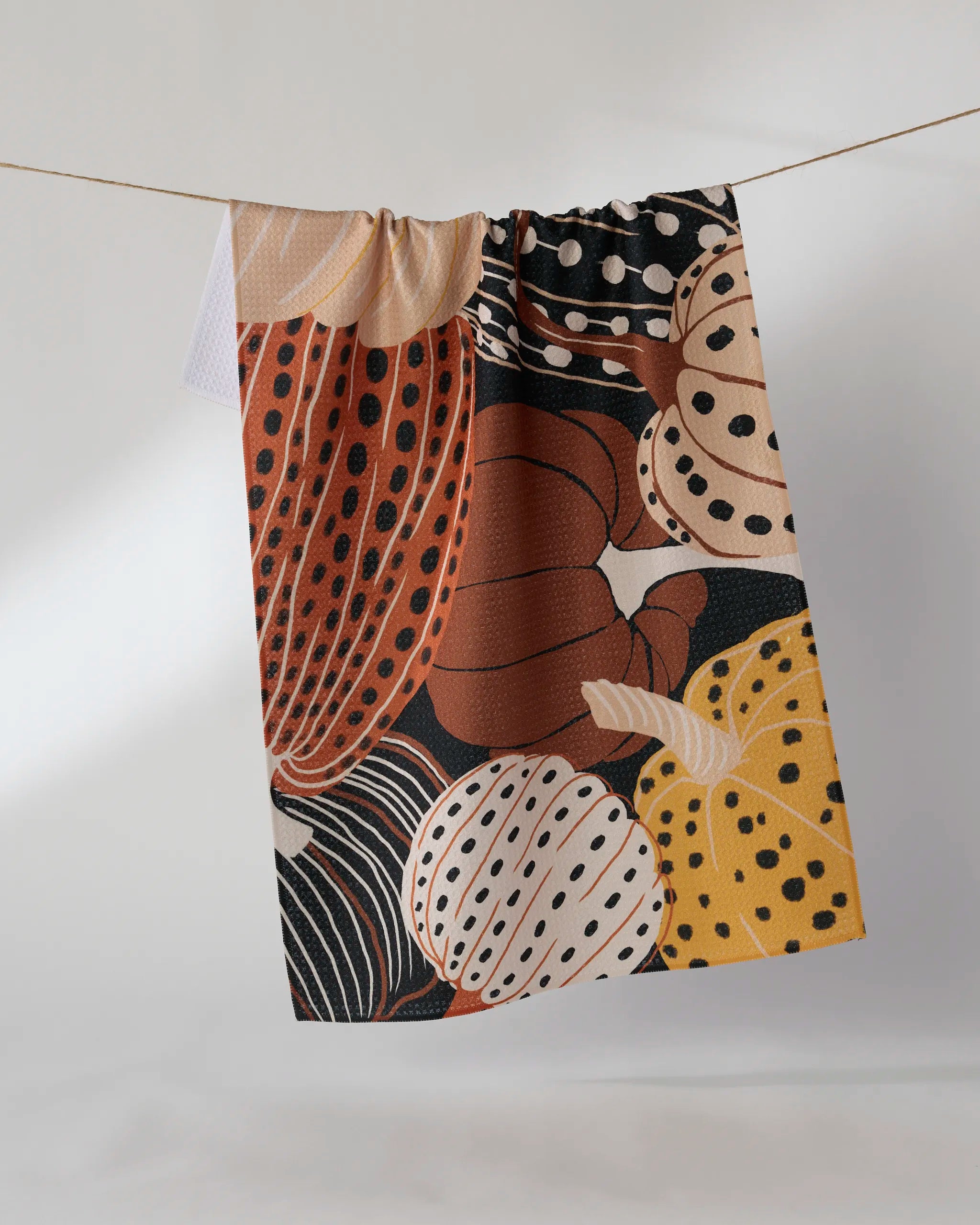 The "Painted Patch" kitchen towel by Geometry, featuring an abstract pattern of spotted and striped gourds in brown, yellow, cream, and orange, hangs on a line against a white background.
