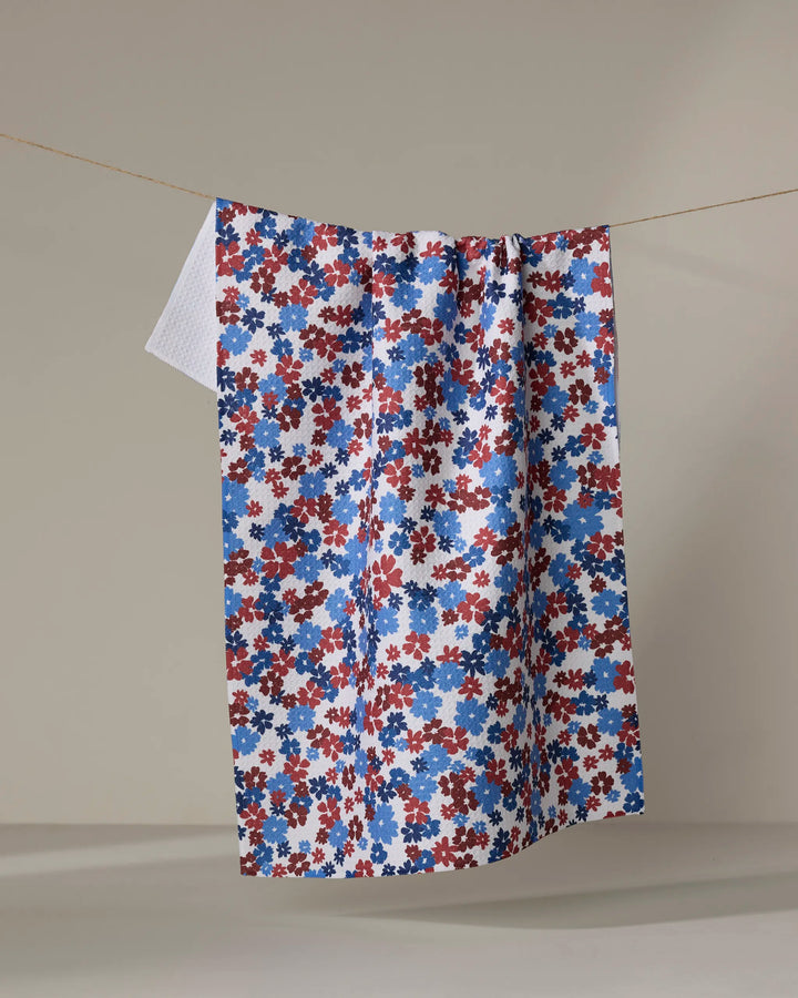 The Ashley Celena Painted Petals ultra-absorbent kitchen towel, featuring a small red, blue, and white floral pattern, hangs on a clothesline against a plain beige background.