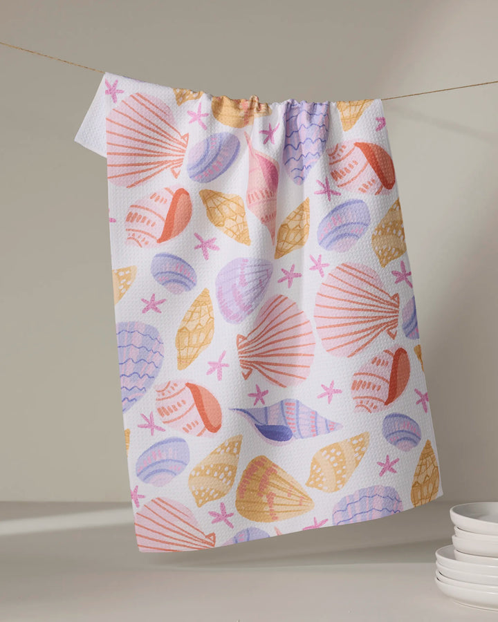 The Painted Shells tea towel by Janna Sue Design hangs on a line, showing off vibrant seashell and starfish prints. Made from quick-drying recycled materials, it brightens the scene while white plates rest in the lower right corner.