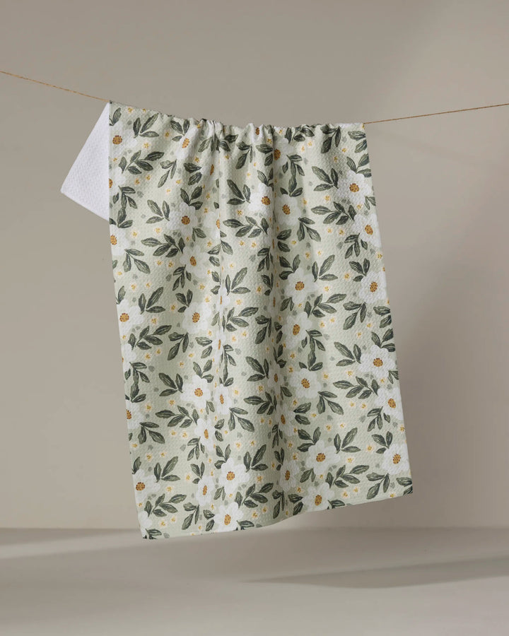 The "Painted Spring Flowers" tea towel by Teri the Butter features green leaves and white daisies with yellow centers on a white background, hanging on a string against a neutral backdrop.