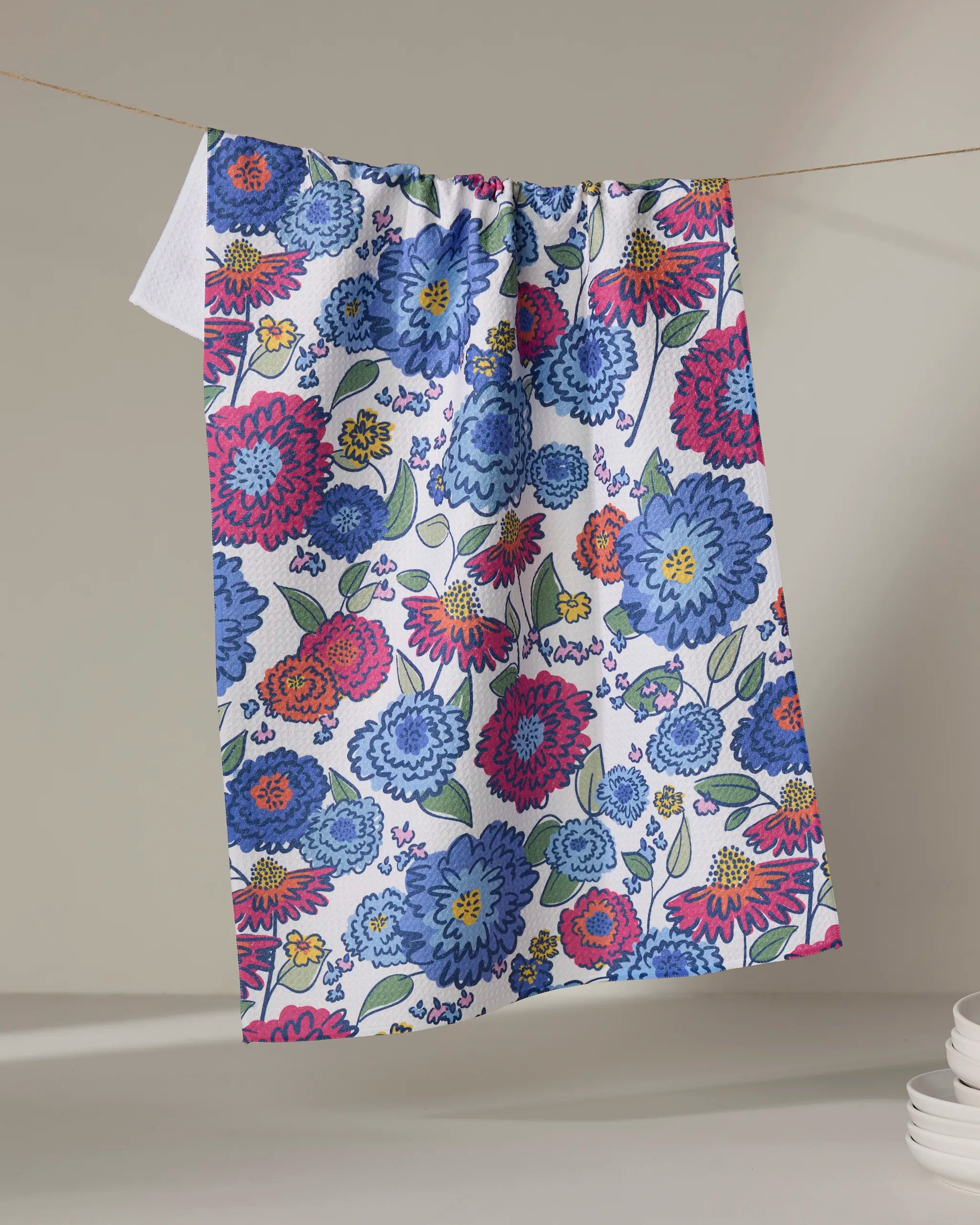 The Painterly Meadow tea towel by Kris Ruff features a vibrant pattern of blue, red, and pink flowers with green leaves, crafted from recycled materials. It hangs on a neutral background with stacked white dishes nearby.