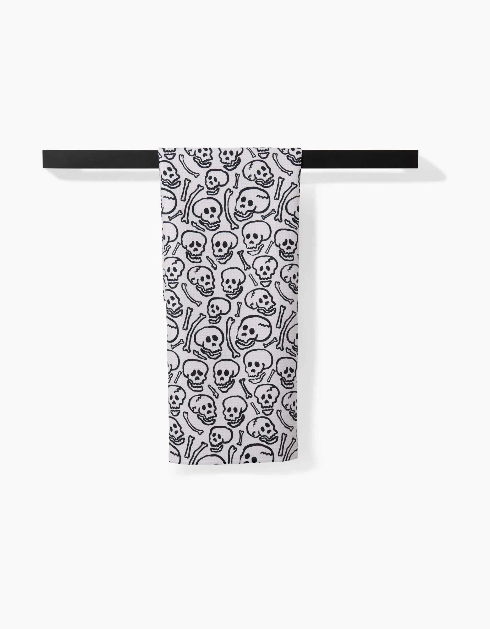 The Kayla Ann Painterly Skulls kitchen towel, made from white recycled material and featuring a repeating black skull and crossbones pattern, hangs neatly over a simple black bar against a white background.