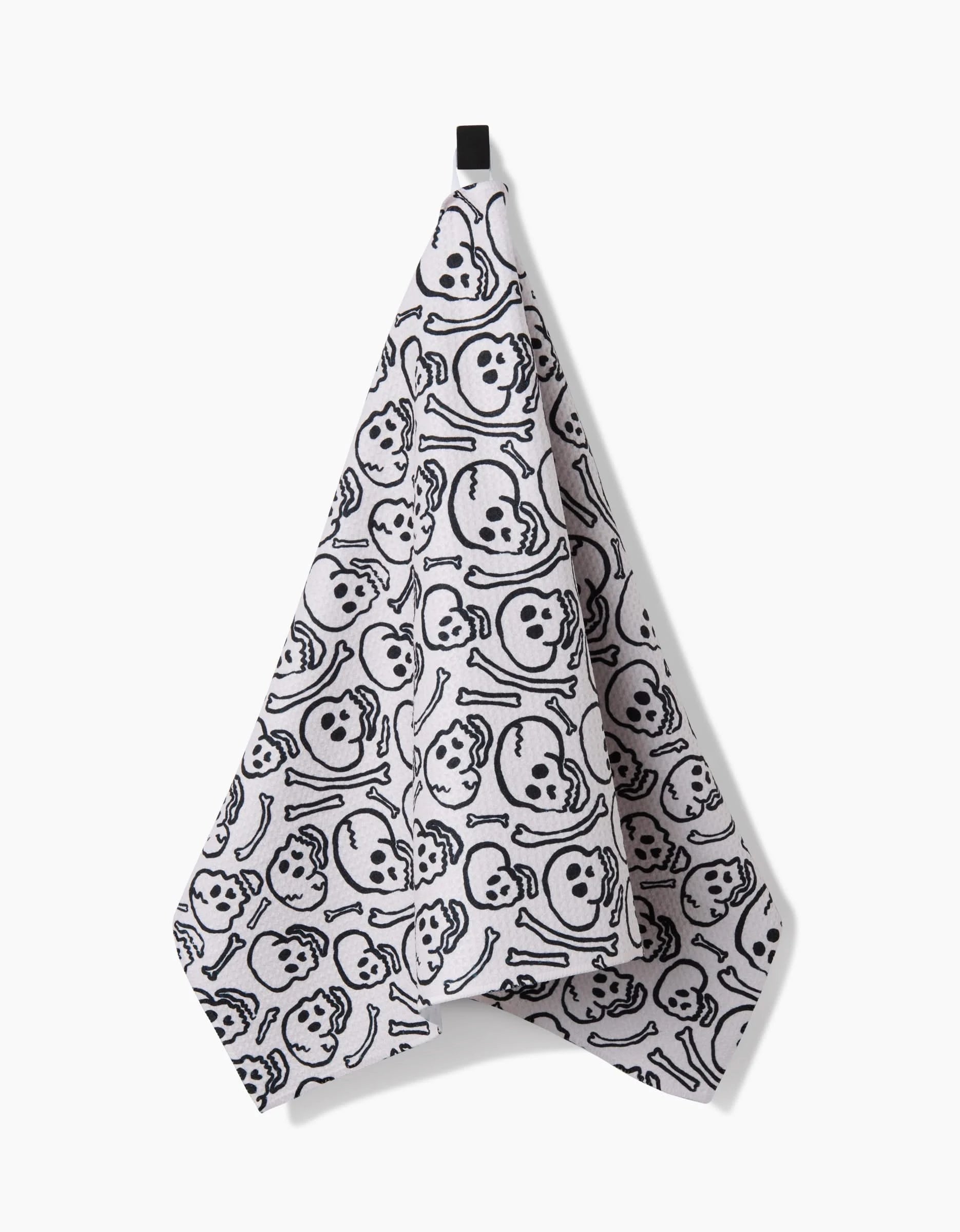 A Kayla Ann Painterly Skulls tea towel features black skulls and crossbones on ultra-absorbent Geoweave™ fabric, hanging from a black loop against a white background.