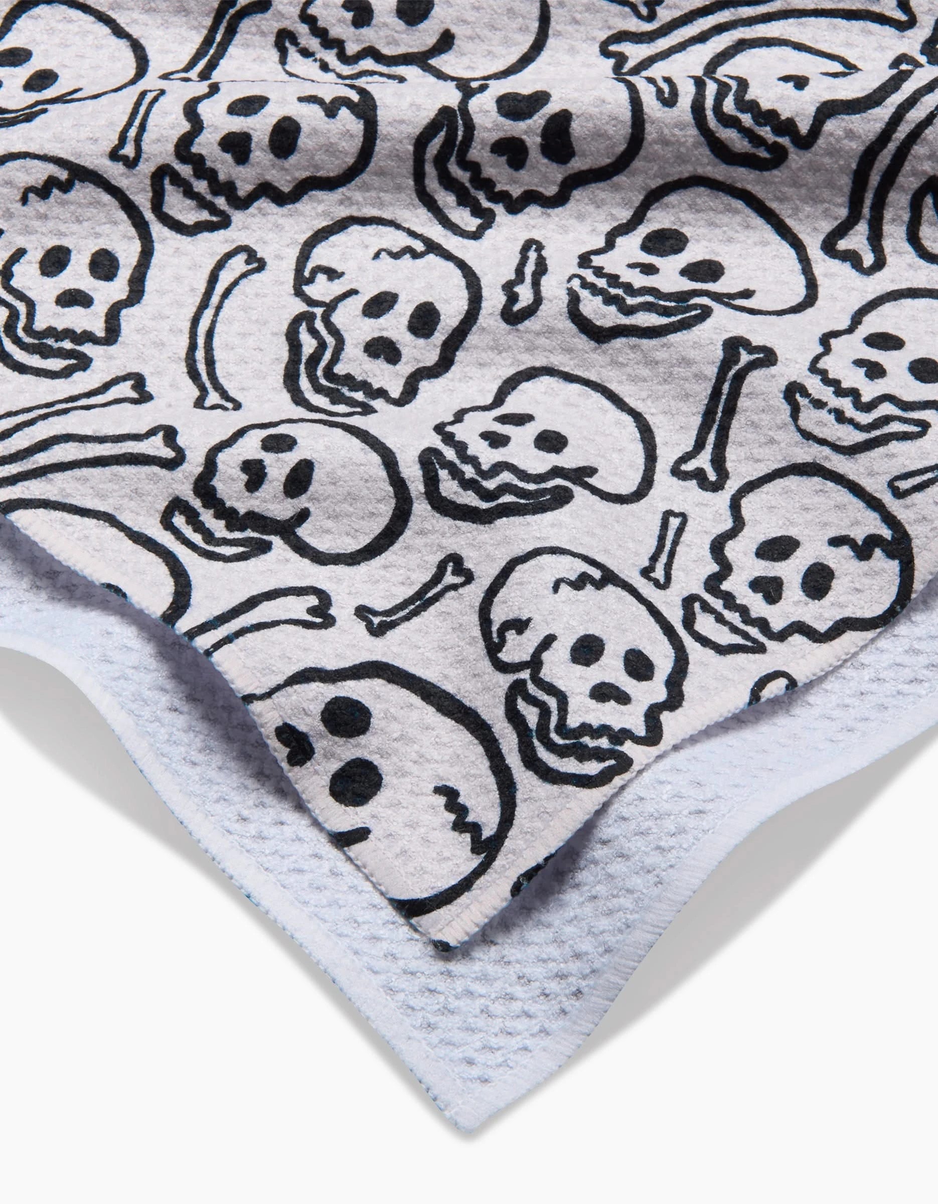 A close-up of the Kayla Ann Painterly Skulls Tea Towel, showing a black cartoon skull and bones pattern on ultra-absorbent Geoweave™ fabric, overlapping slightly on a white textured surface.