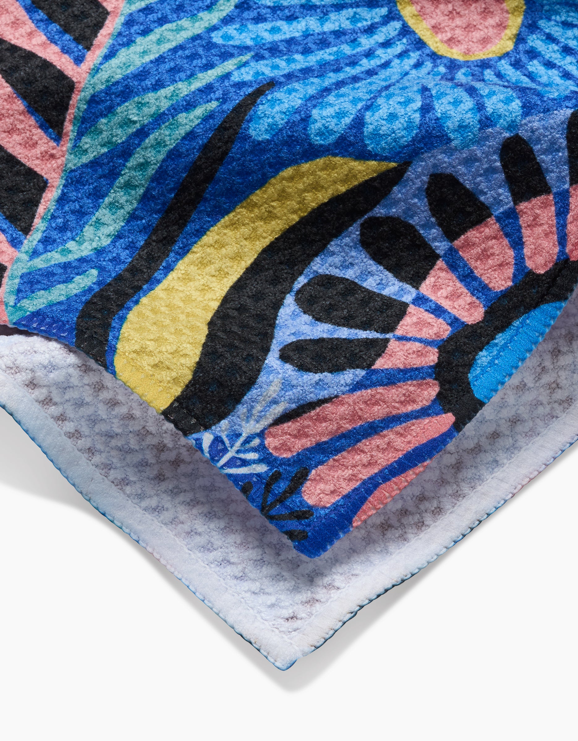 A close-up of the Palm Beach tea towel by Masha Volnova shows its blue, yellow, black, and pink abstract floral design on a textured Geoweave™ surface with stitched edges and a white recycled-material underside.