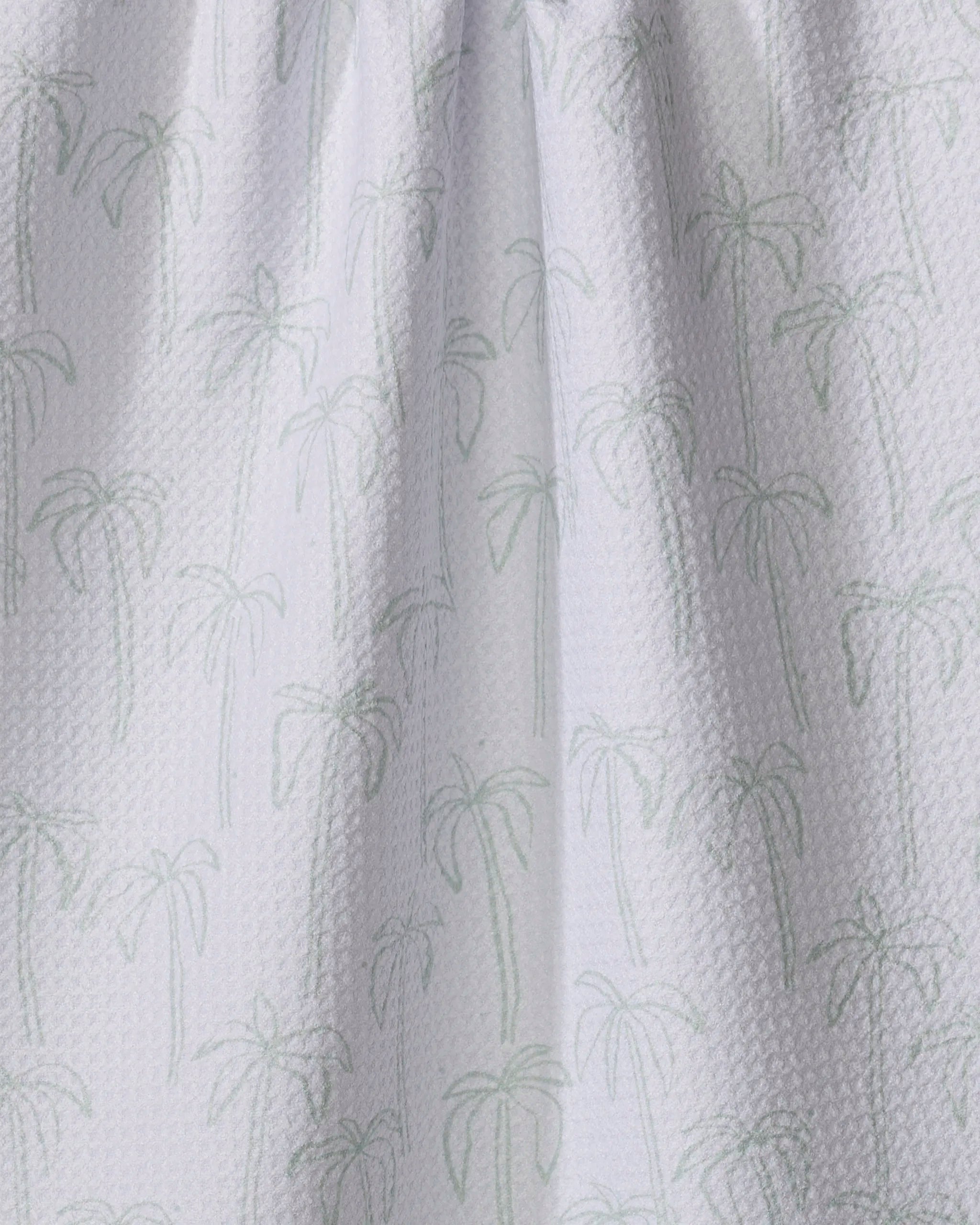 The Palms tea towel by Jen B. Peters features a white textured fabric with a subtle light green palm tree pattern. Made from quick-drying recycled materials, its gentle folds create soft shadows for a stylish, eco-friendly look.