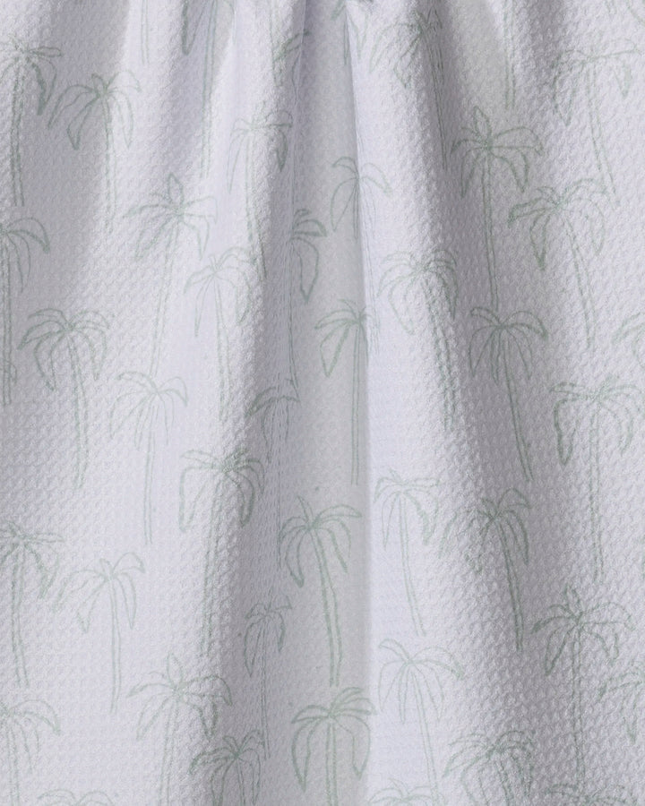 The Palms tea towel by Jen B. Peters features a white textured fabric with a subtle light green palm tree pattern. Made from quick-drying recycled materials, its gentle folds create soft shadows for a stylish, eco-friendly look.