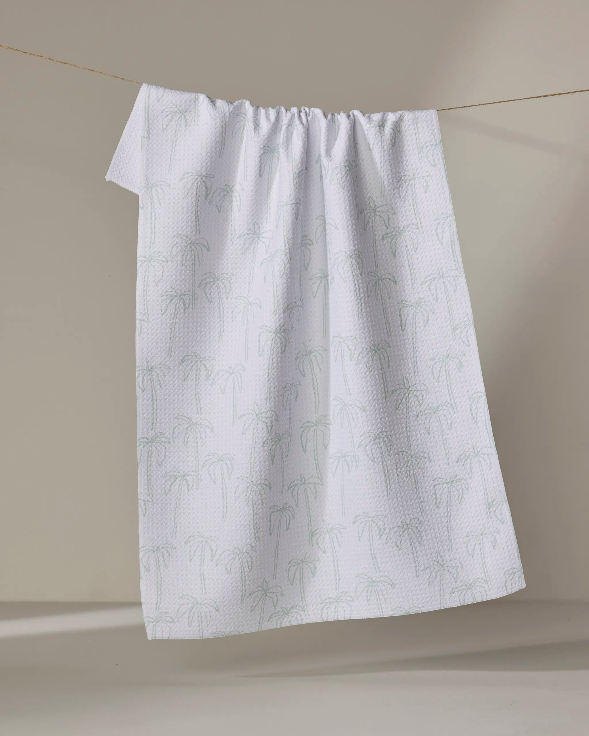 Palms" by Jen B. Peters—a white tea towel featuring a subtle palm tree design and crafted from recycled materials—hangs on a clothesline against a neutral background.