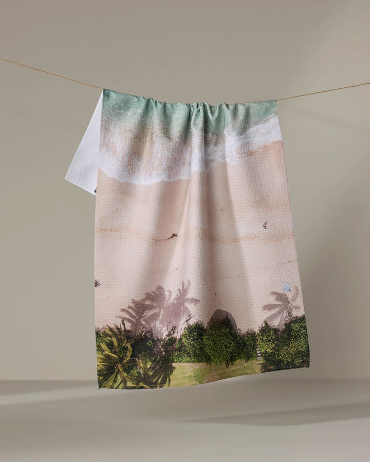 The Kait Hanson "Paradise Found" towel, featuring ocean waves, a sandy shore, and palm trees, dries quickly and hangs on a line against a plain background.