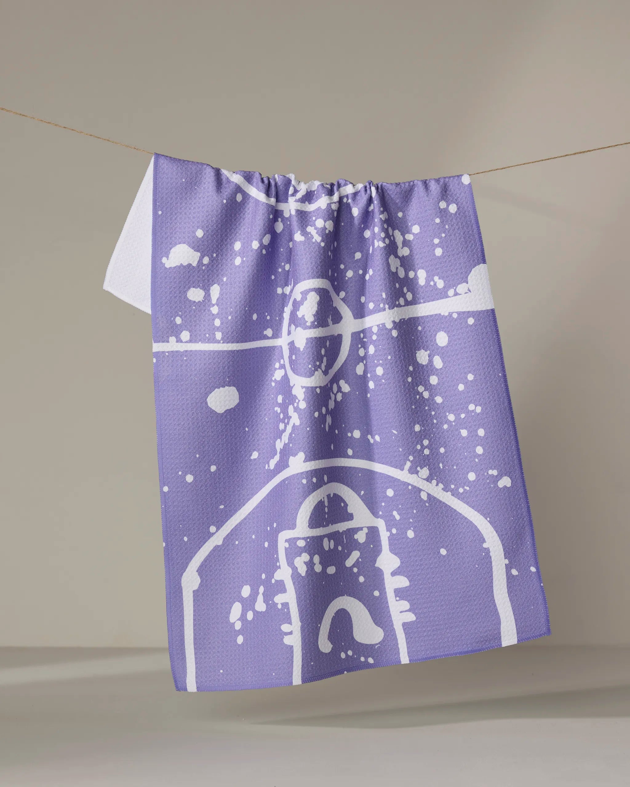 The Sissy Moon "Parquet" kitchen towel, featuring eco-friendly fabric and a purple design with white abstract splatter and lines, hangs on a clothesline against a neutral background.