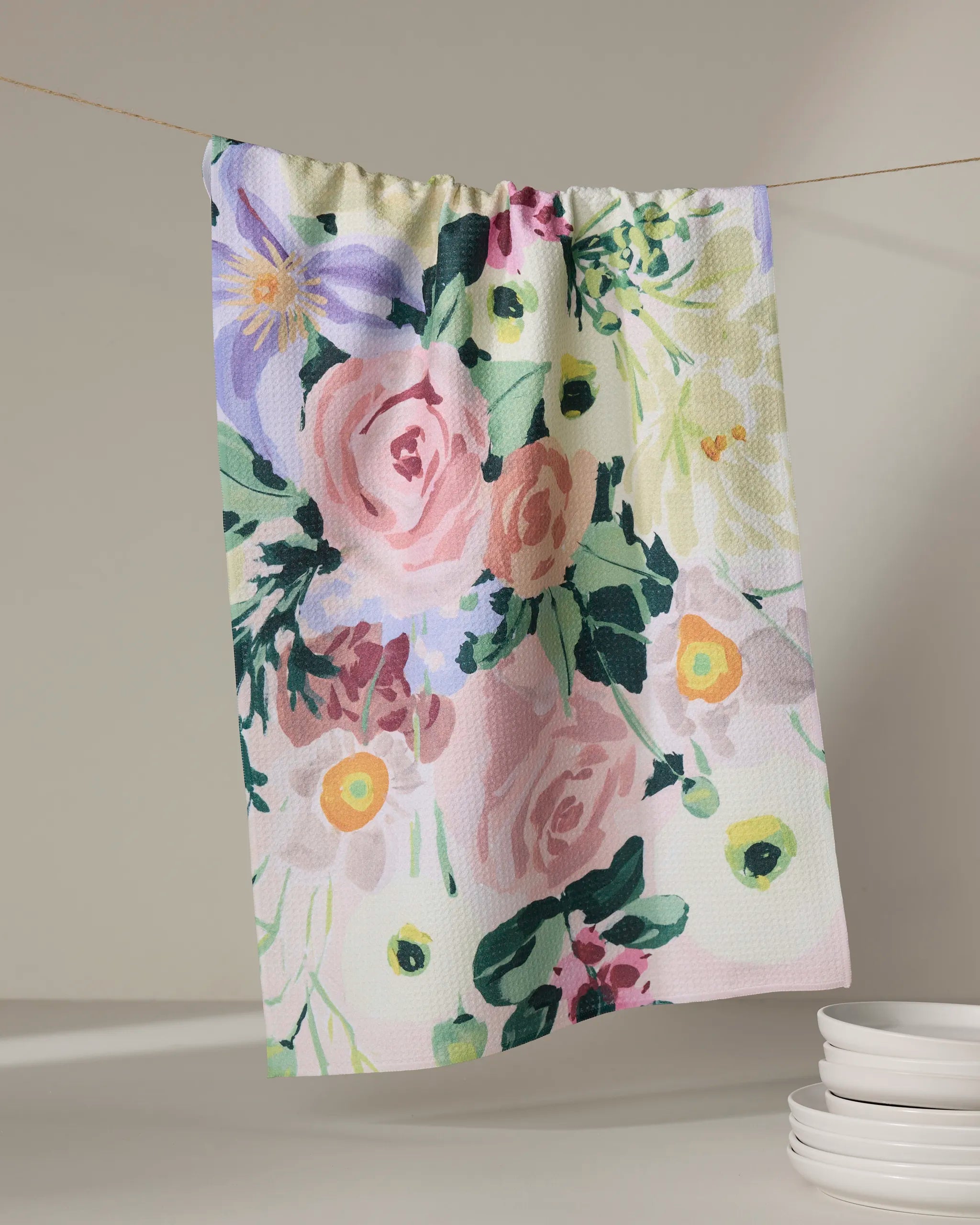 The Pastel Bouquet kitchen towel by Ania Zwara Art, made from recycled materials and decorated with pink, purple, and yellow flowers, hangs on a string above a white counter with stacked plates nearby.