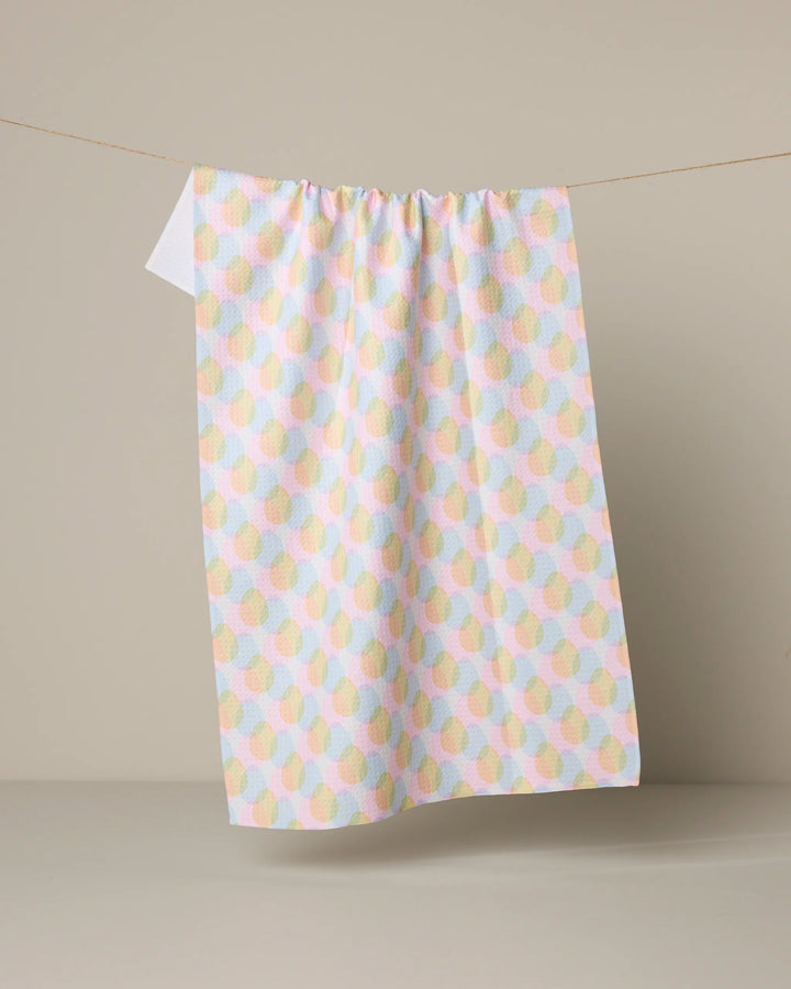 The "Pastel Pop" tea towel by Geometry, made from recycled materials and featuring overlapping circles in pastel hues, hangs on a string against a neutral beige background.
