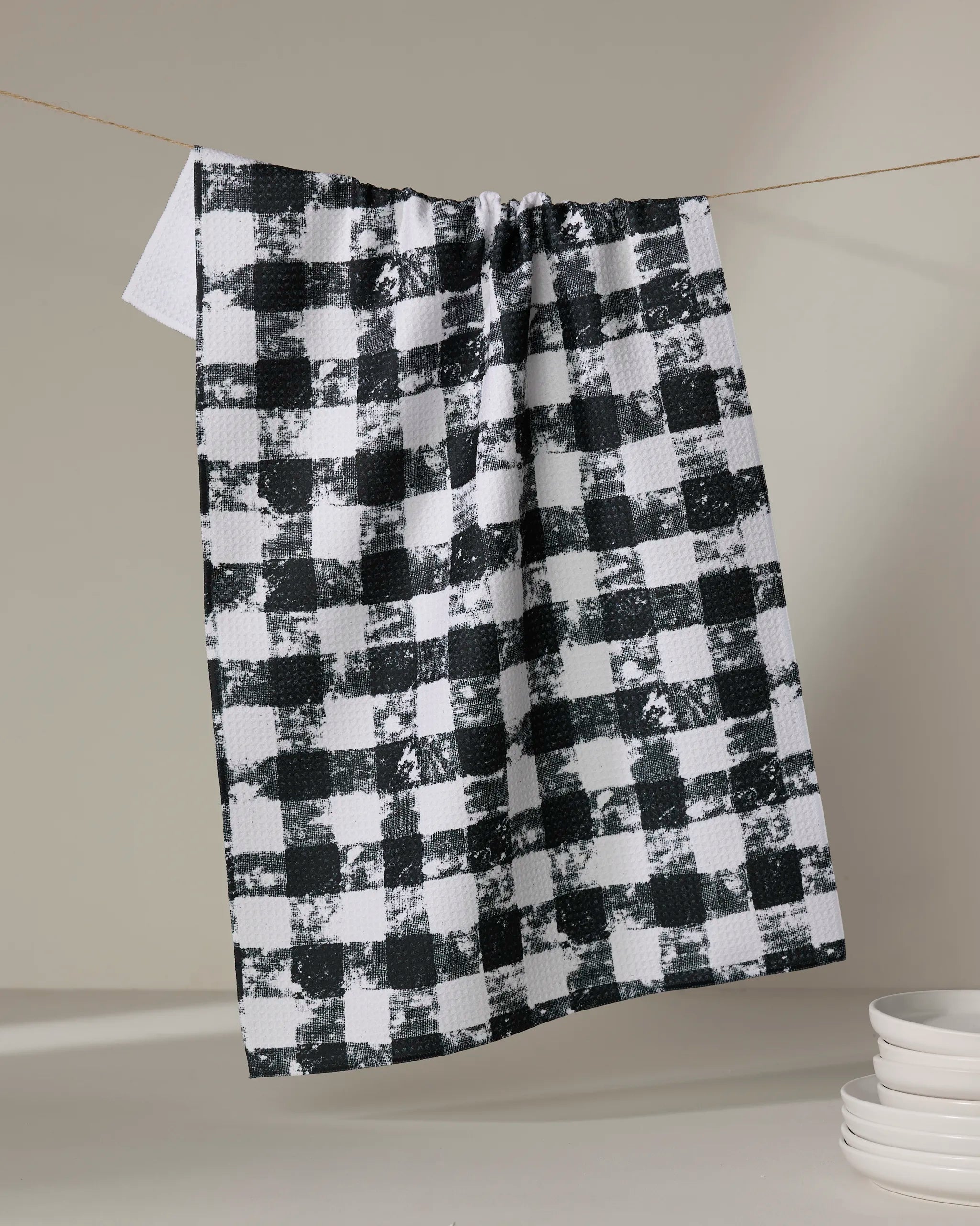 The Patch Ash recycled kitchen towel by Geometry in a black and white checkered pattern hangs on a string against a neutral background, with white dishes stacked in the lower right corner.
