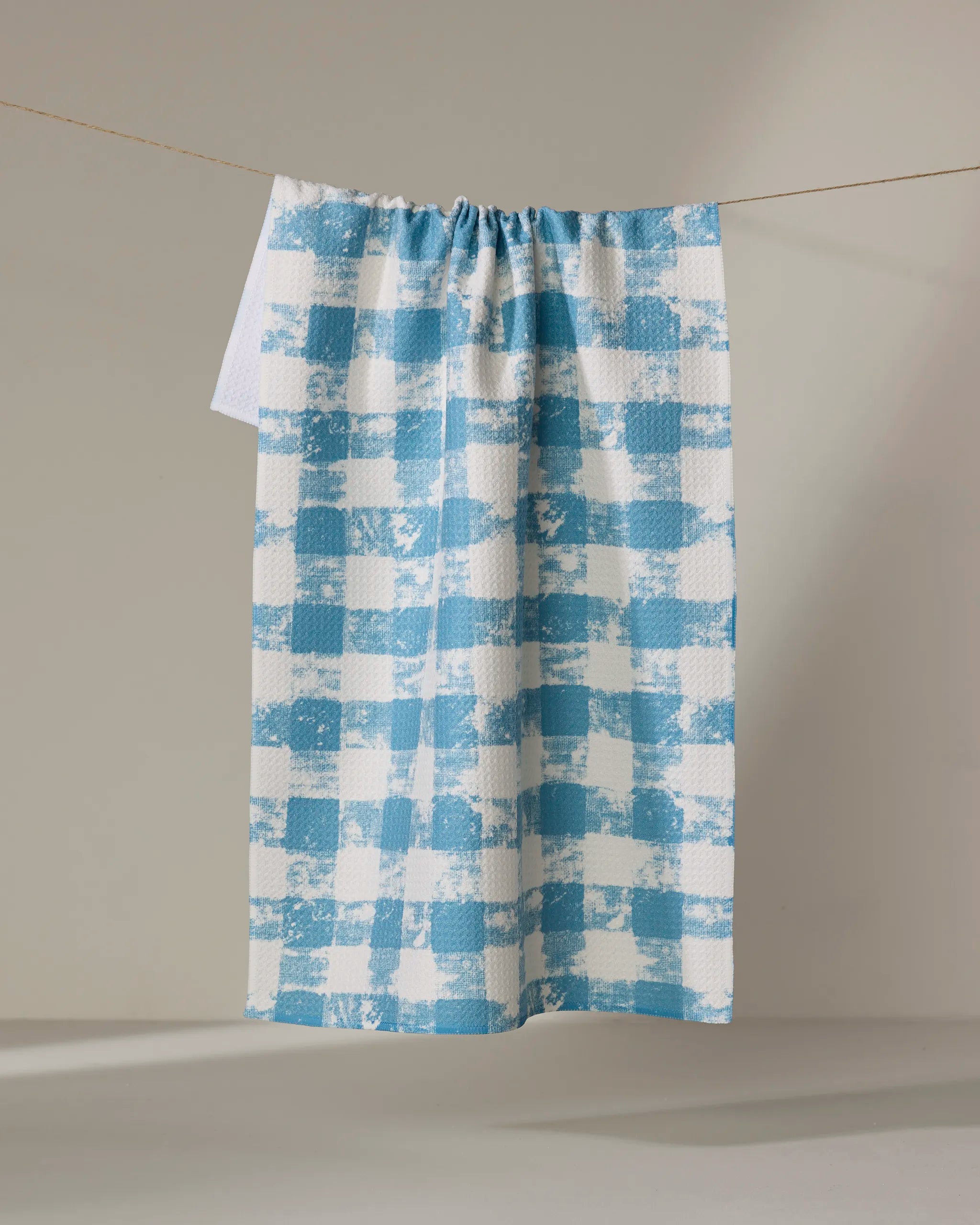 The Geometry Patch Denim recycled kitchen towel, featuring a blue and white checkered textured pattern, hangs on a clothesline against a neutral background.