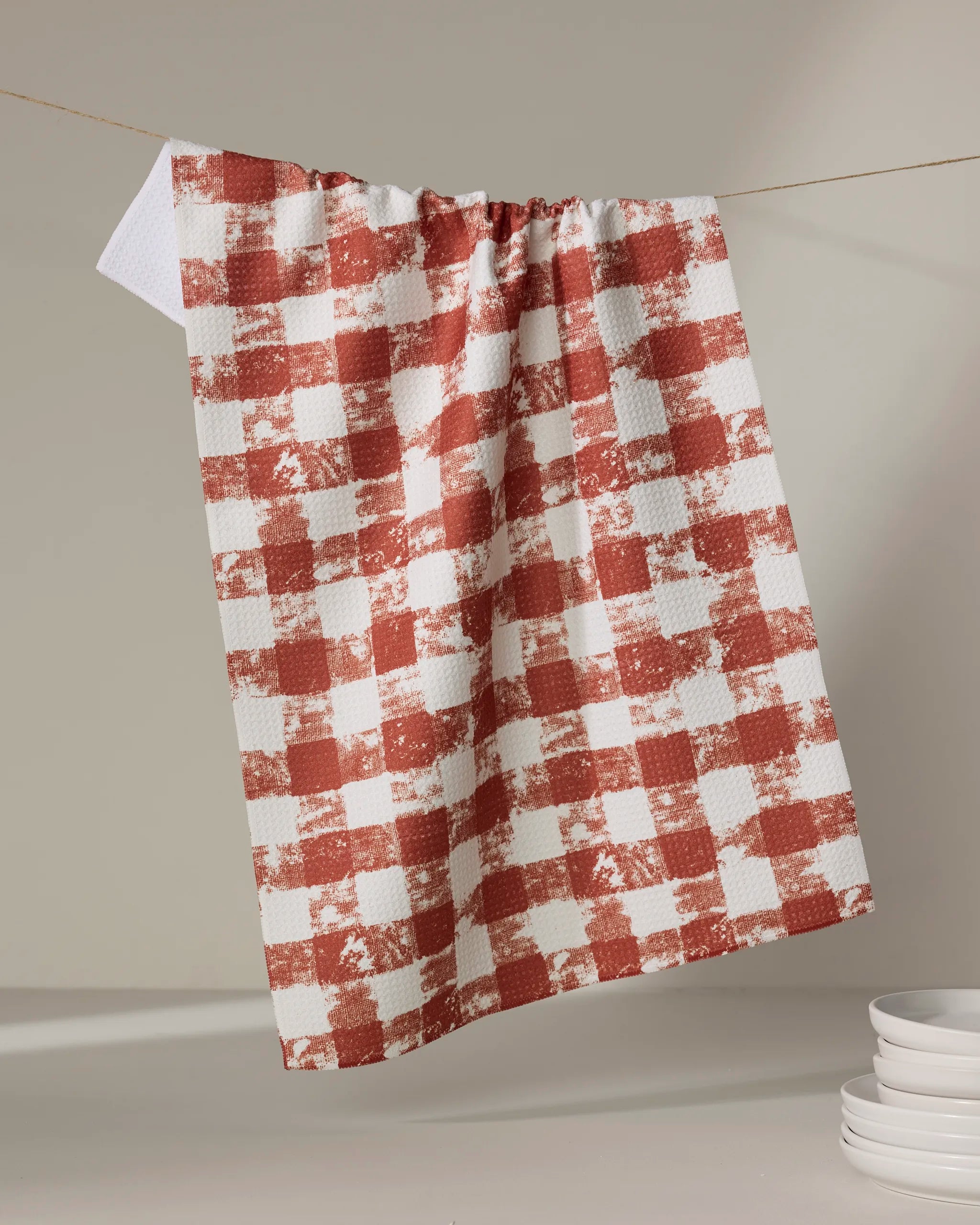 The Geometry Patch Spice tea towel, featuring a red and white checkered design, hangs on a string against a neutral background, with a stack of white plates partially visible in the lower right corner.